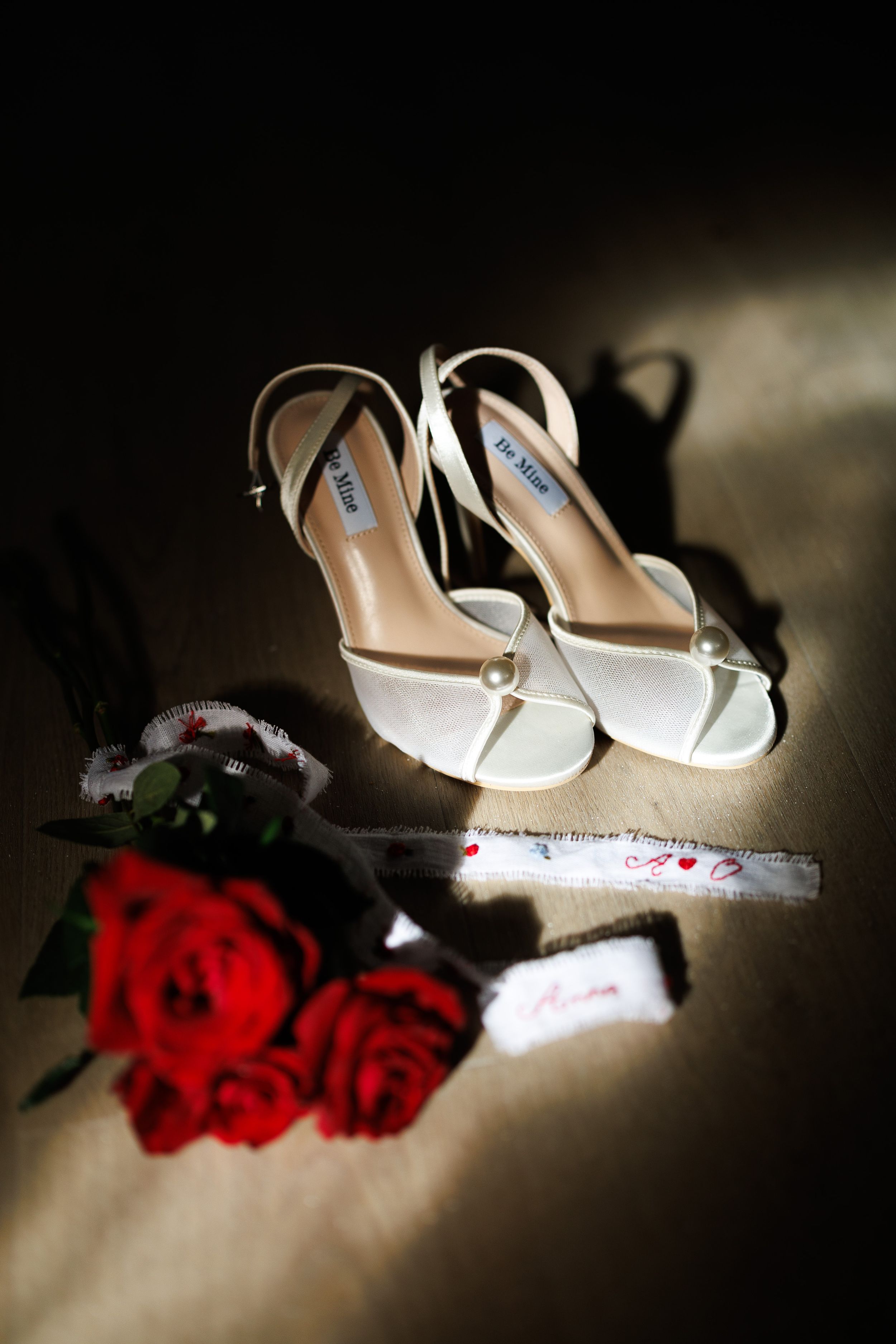 White bridal heels by De Alina displayed with red roses and a wedding garter on a dark surface.