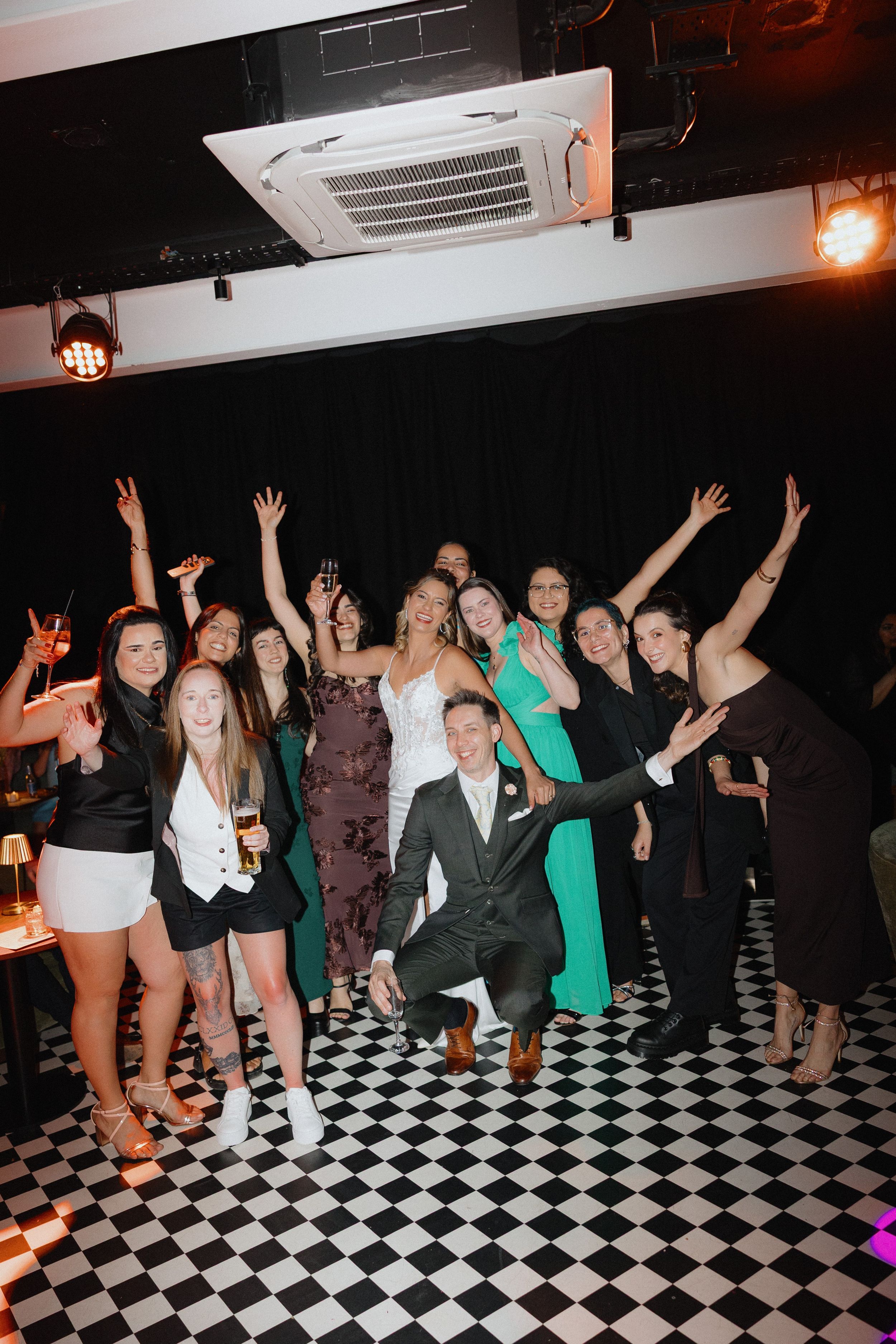 Wedding couple poses with group of friends on checkered dance floor at lively reception venue.
