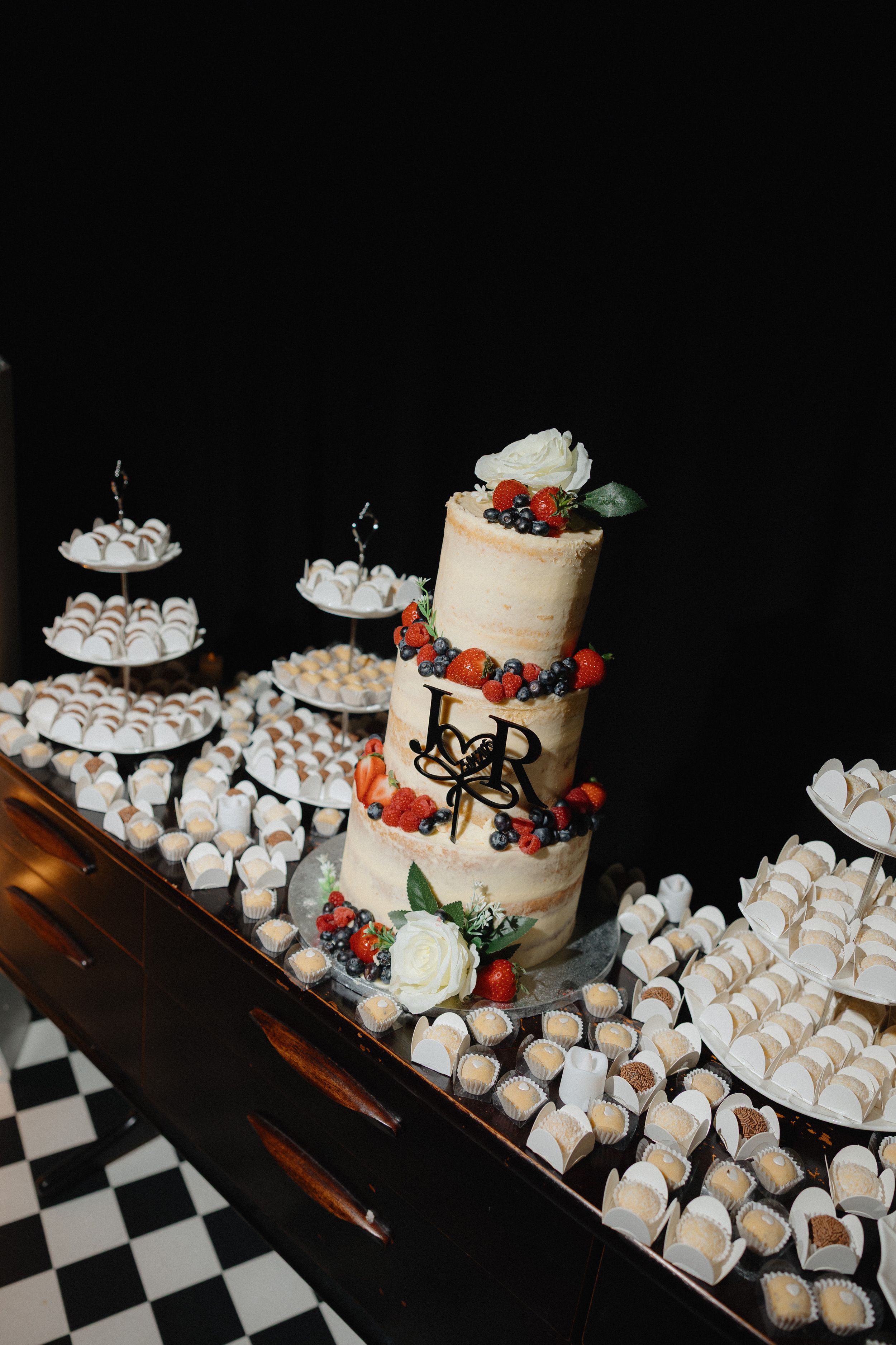 Elegant wedding dessert table featuring a semi-naked tiered cake with fresh berries, surrounded by macarons and chocolate truffles.