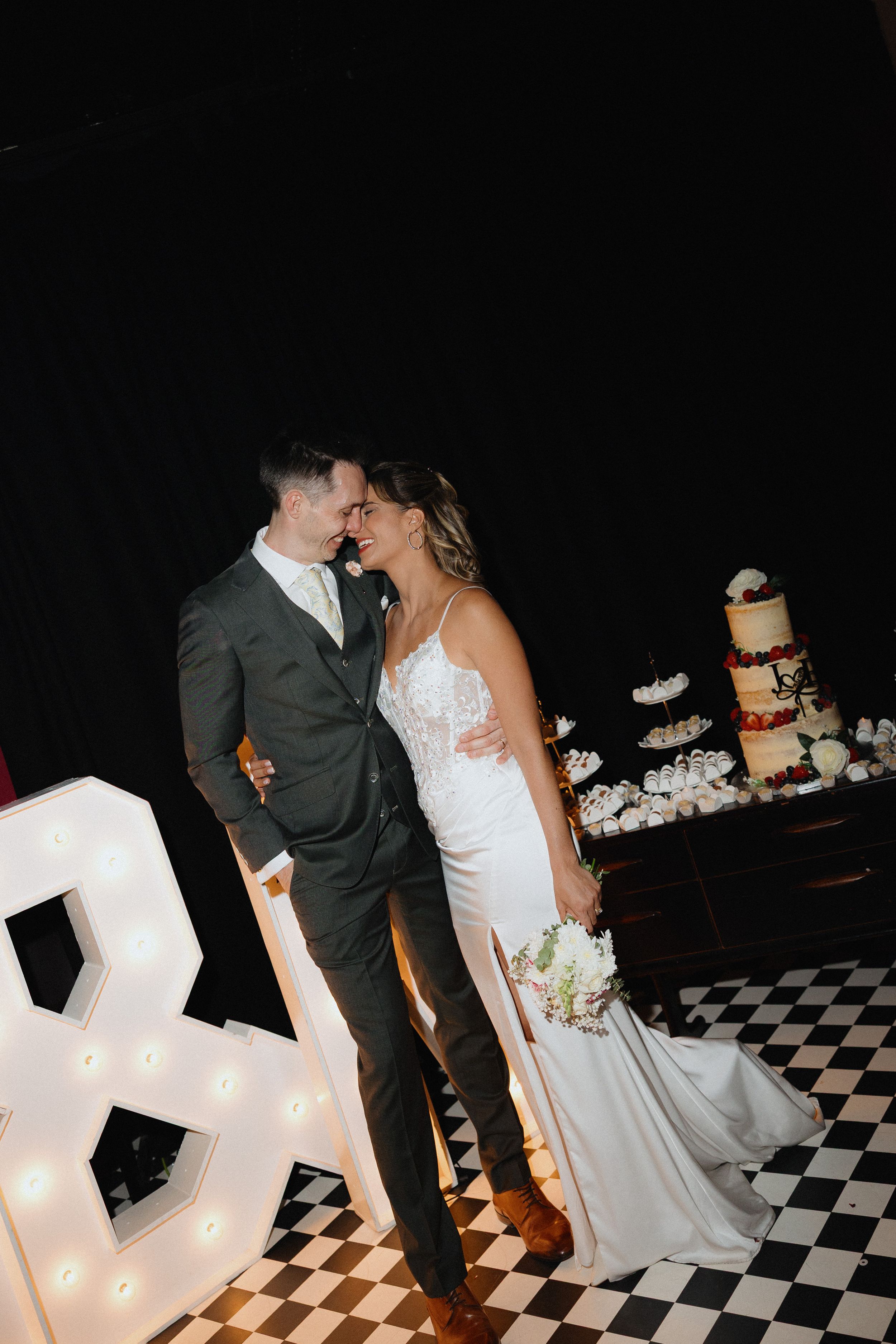 Bride and groom share a romantic moment on a checkered dance floor beside illuminated letters and a wedding cake.