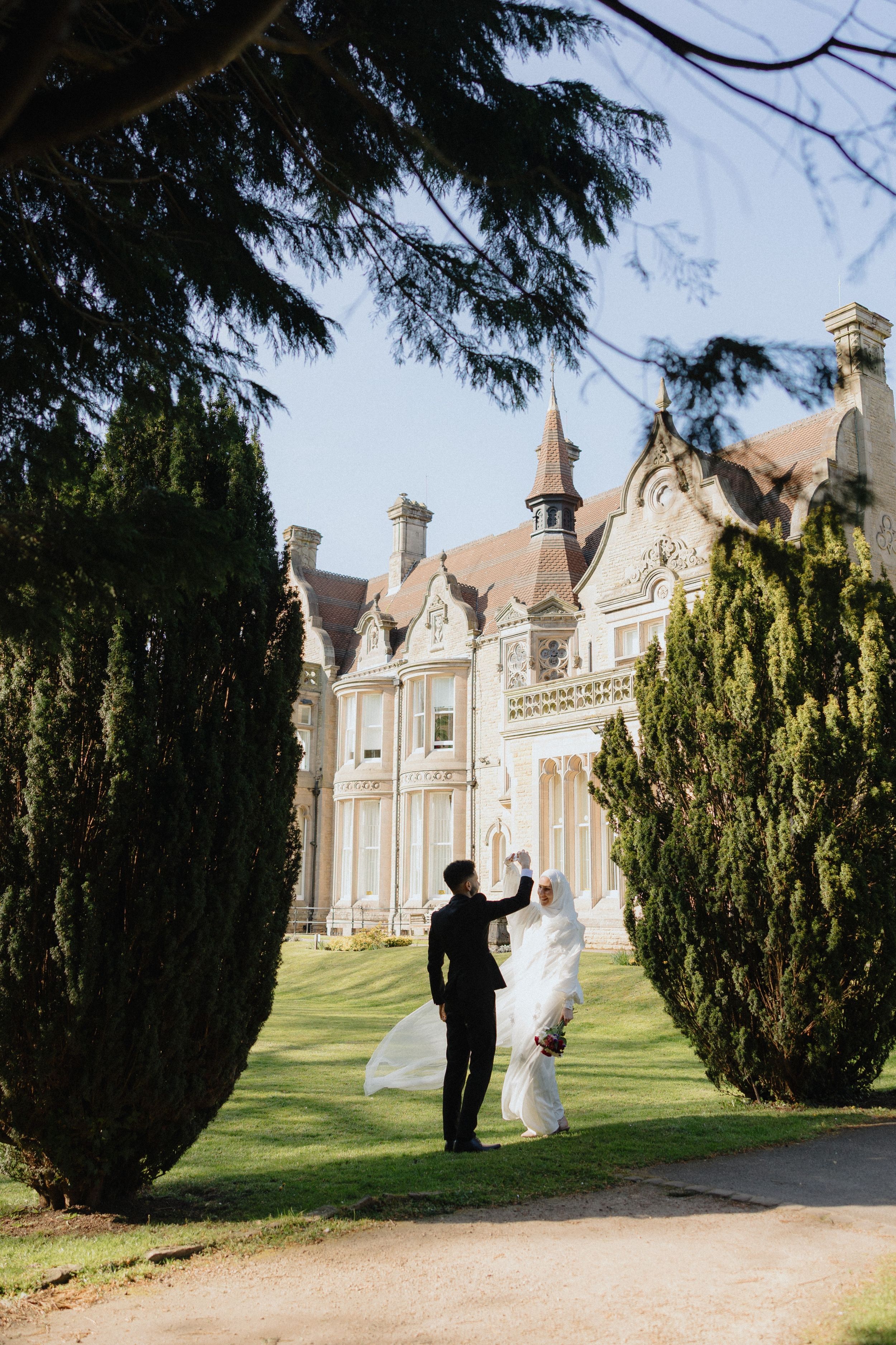 A bride and groom walk hand-in-hand on the grounds of an elegant manor house, framed by tall cypress trees.