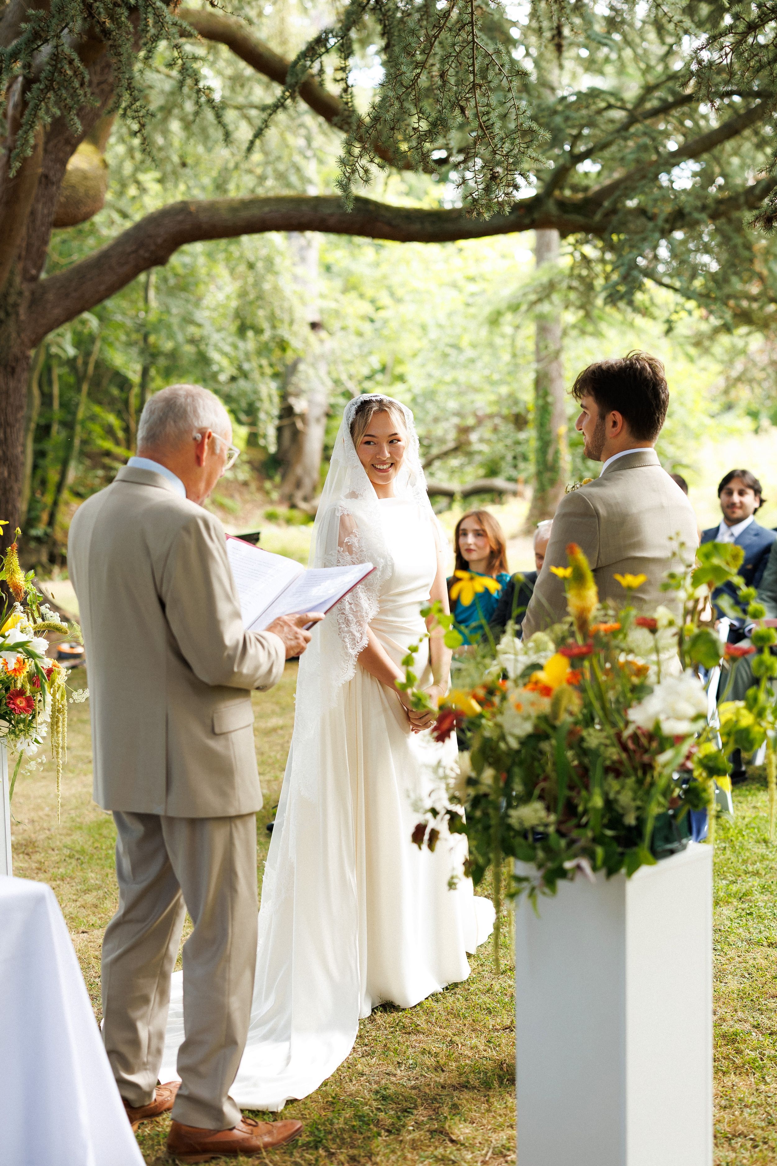 Outdoor wedding ceremony with bride in white gown and veil, groom, and officiant under oak tree with colorful floral arrangements.