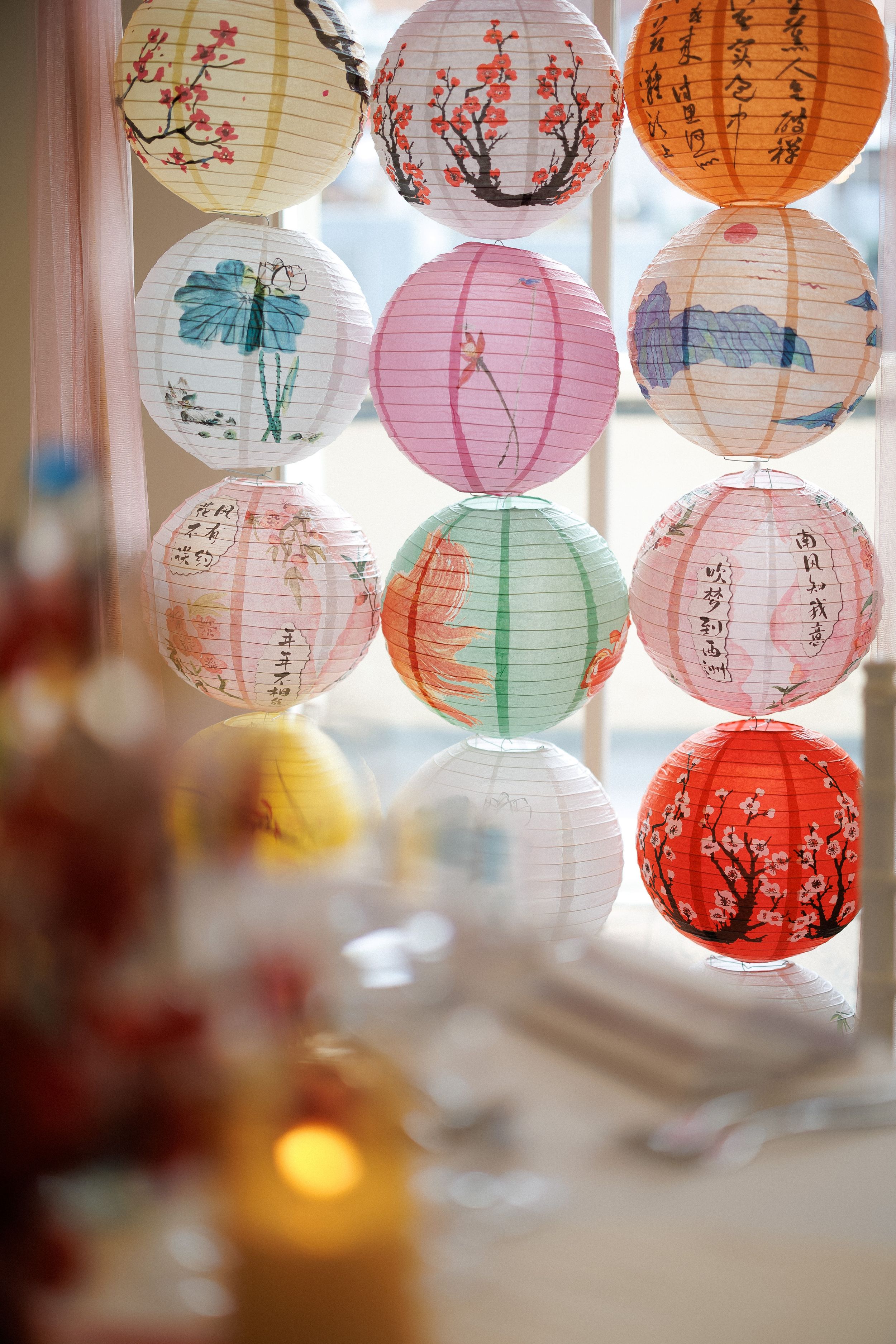 Colorful Chinese paper lanterns with painted floral and calligraphy designs hanging in rows by a bright window.