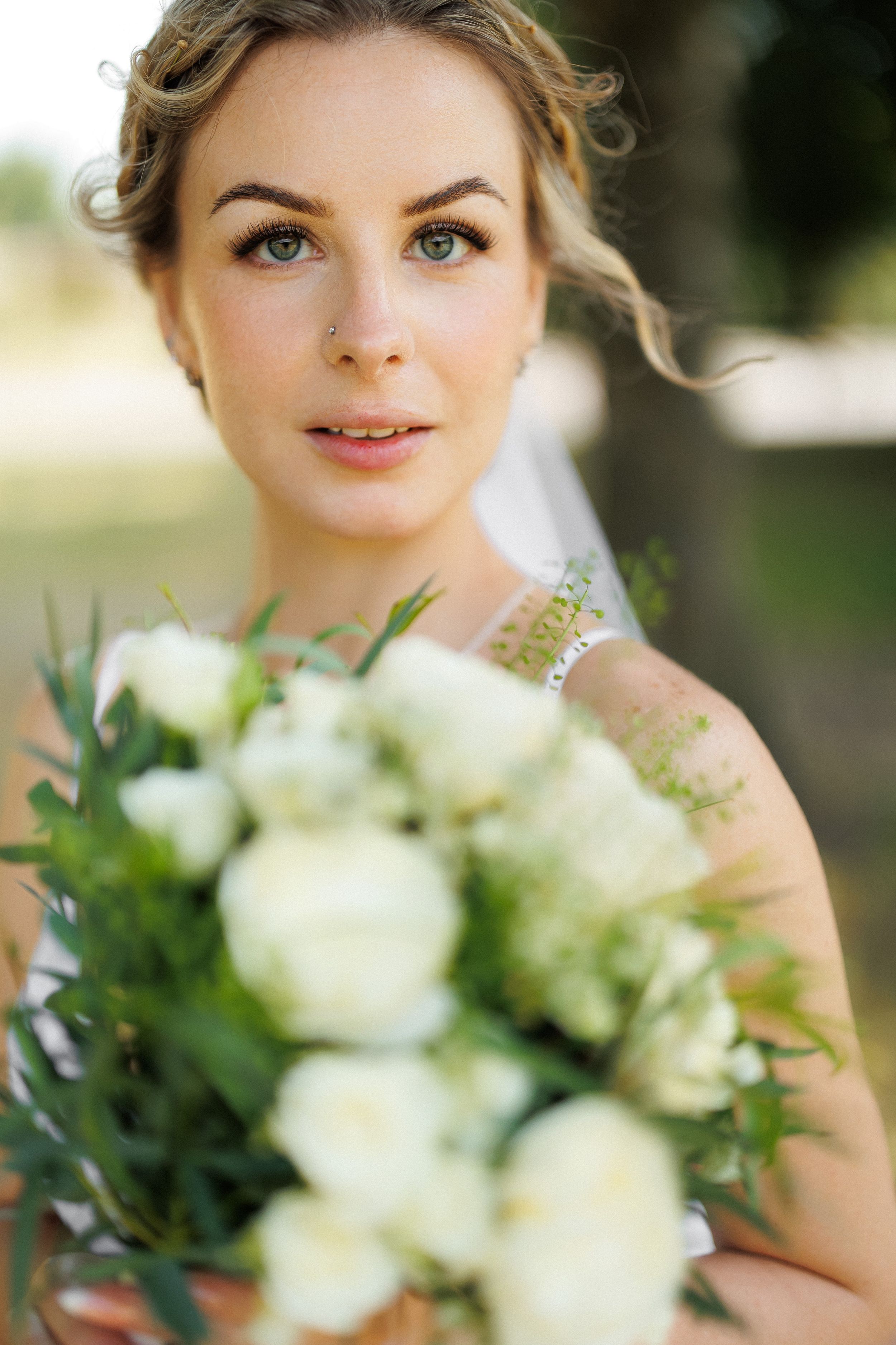 A beautiful bride holds a white rose bouquet with greenery outdoors, wearing a veil and natural makeup.