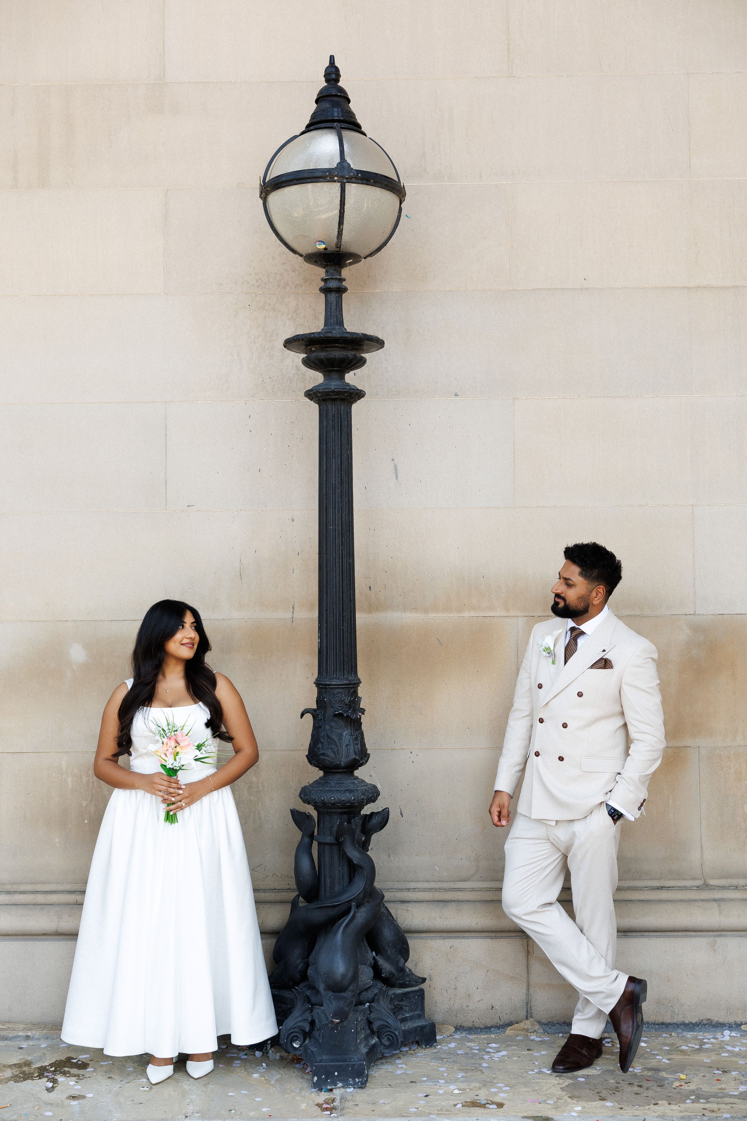 Bride in white gown and groom in cream suit pose by ornate black lamppost against stone wall.