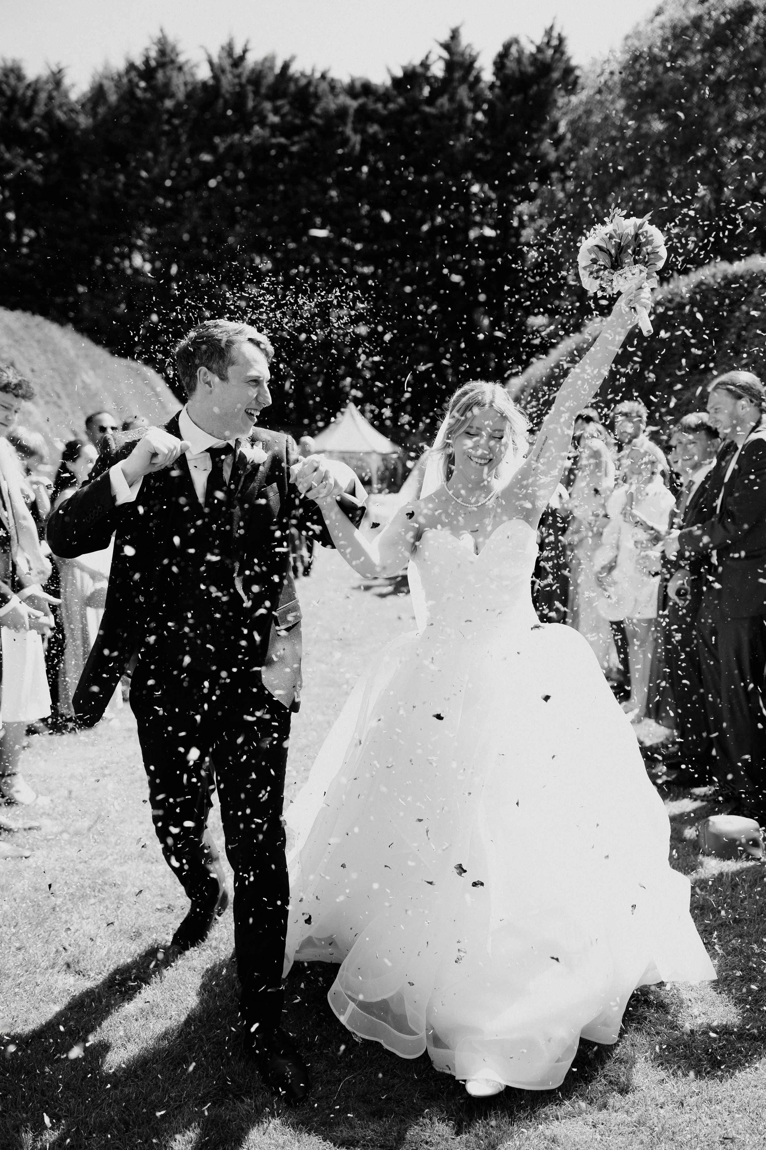 Black and white photo of joyful bride and groom walking through confetti shower after wedding ceremony.