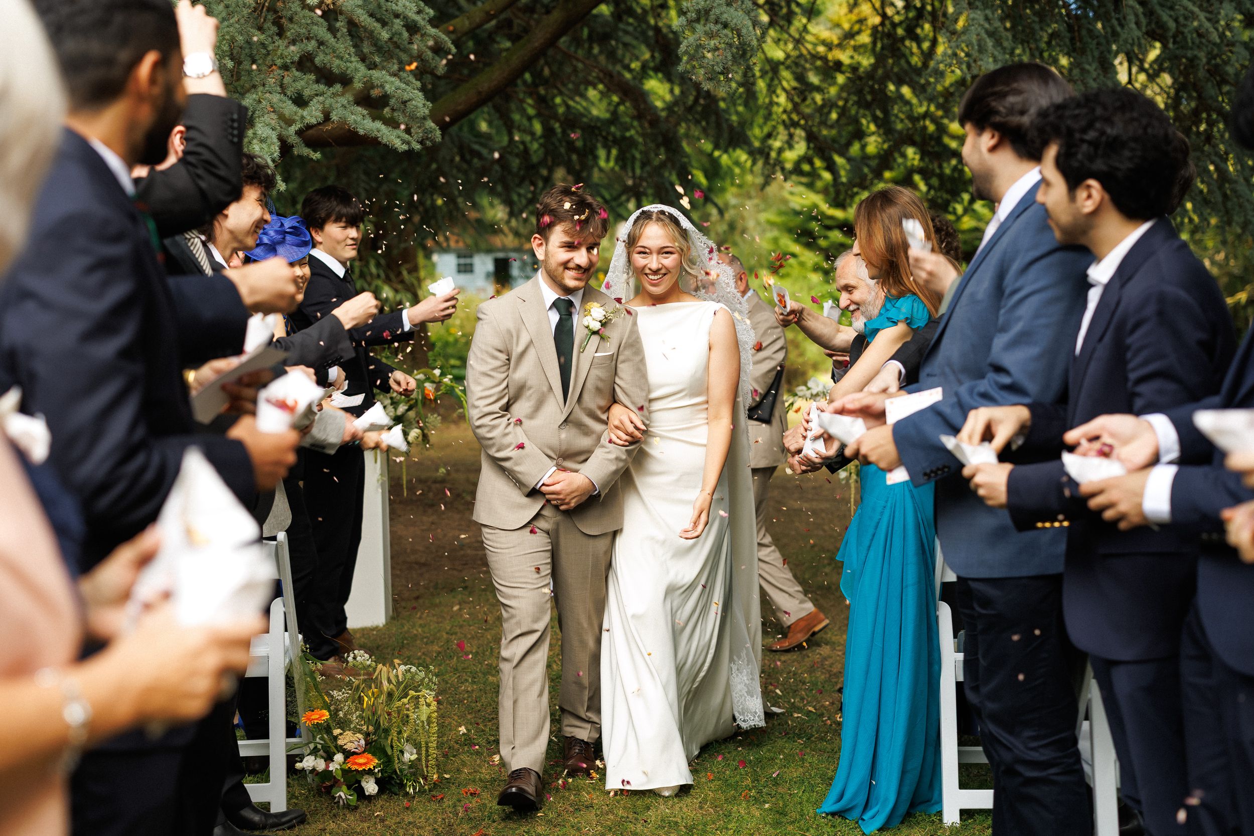 Bride and groom walk down outdoor aisle as guests toss confetti at a garden wedding ceremony.