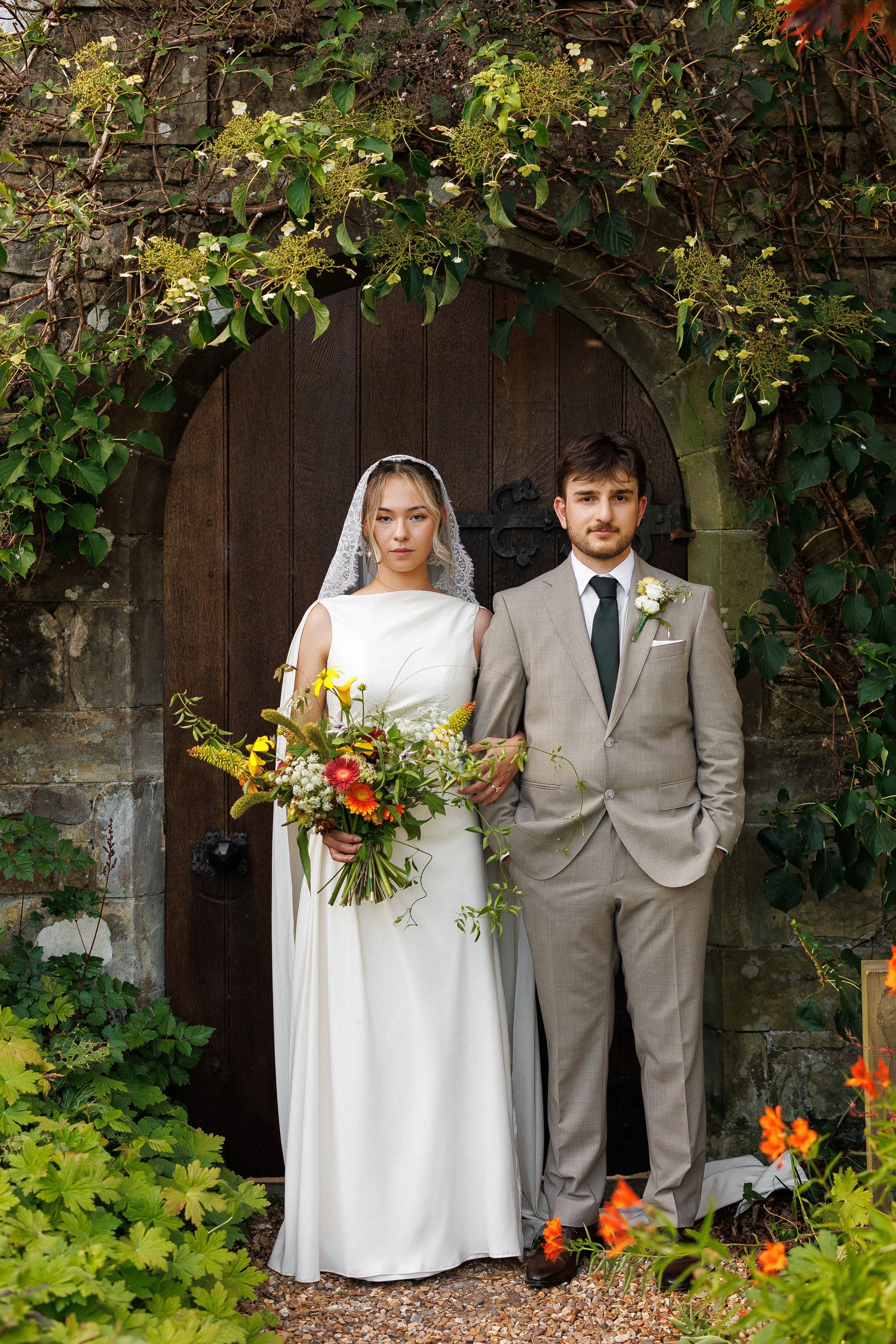 Bride in white gown with veil holding colorful bouquet stands beside groom in gray suit before rustic wooden door.