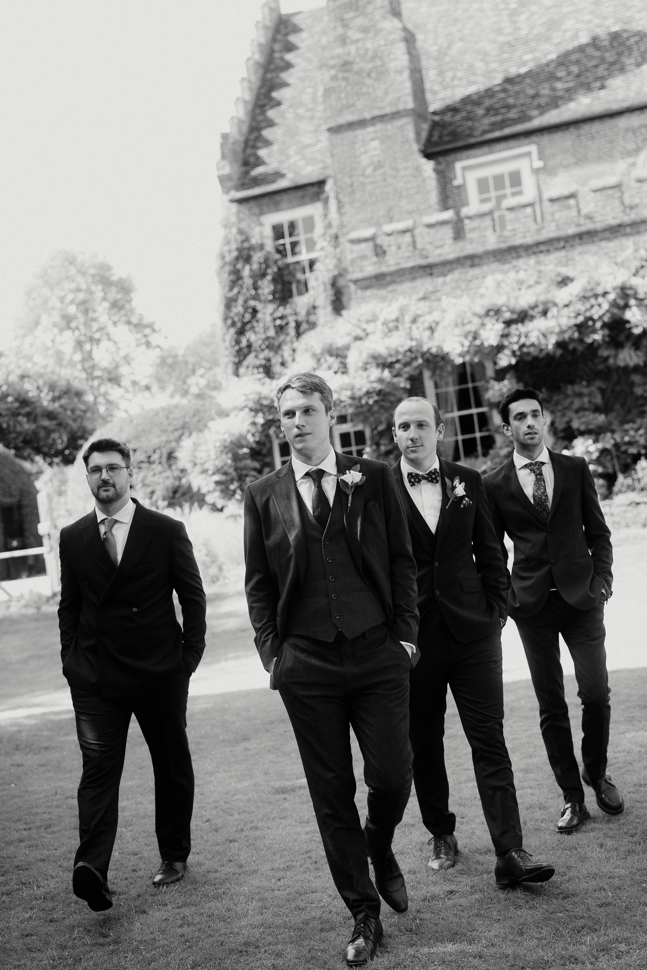 Four groomsmen in dark suits pose outdoors near a historic stone building in this elegant black and white wedding photo.