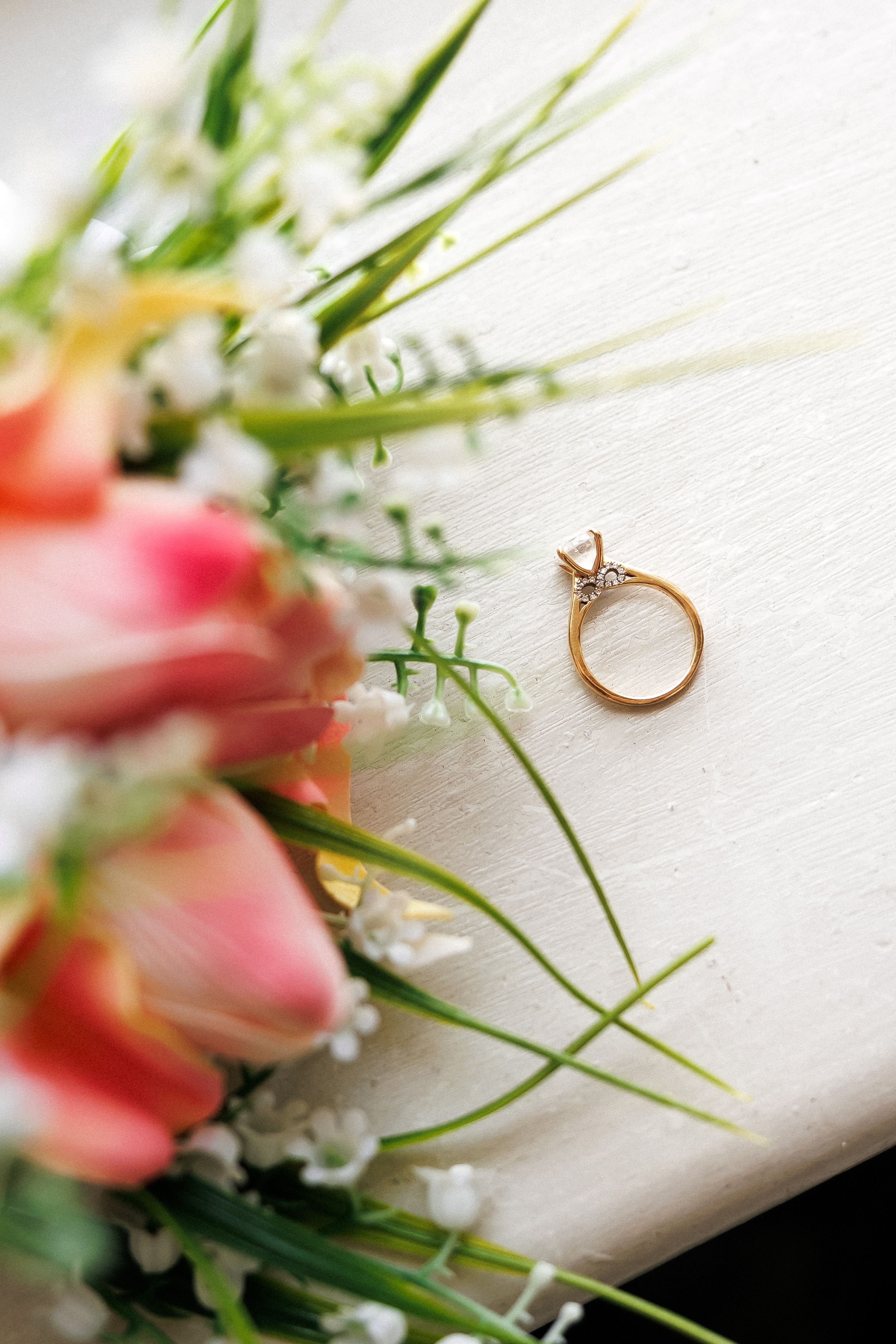 Gold diamond engagement ring resting on white surface beside a bouquet of pink tulips and baby's breath flowers.