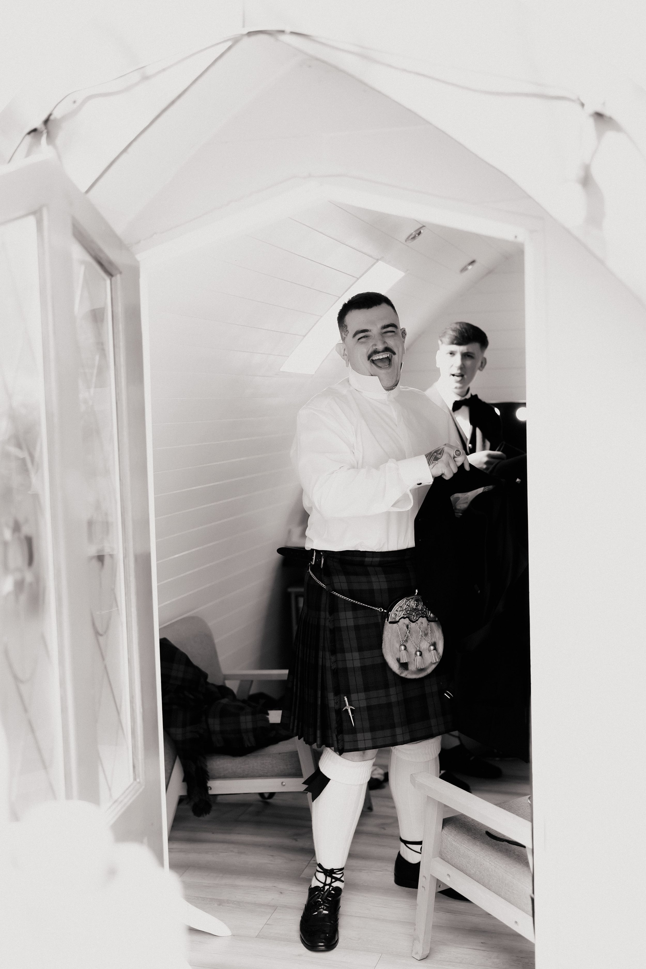 Smiling groom in traditional Scottish kilt and white shirt stands in doorway with groomsman behind him.