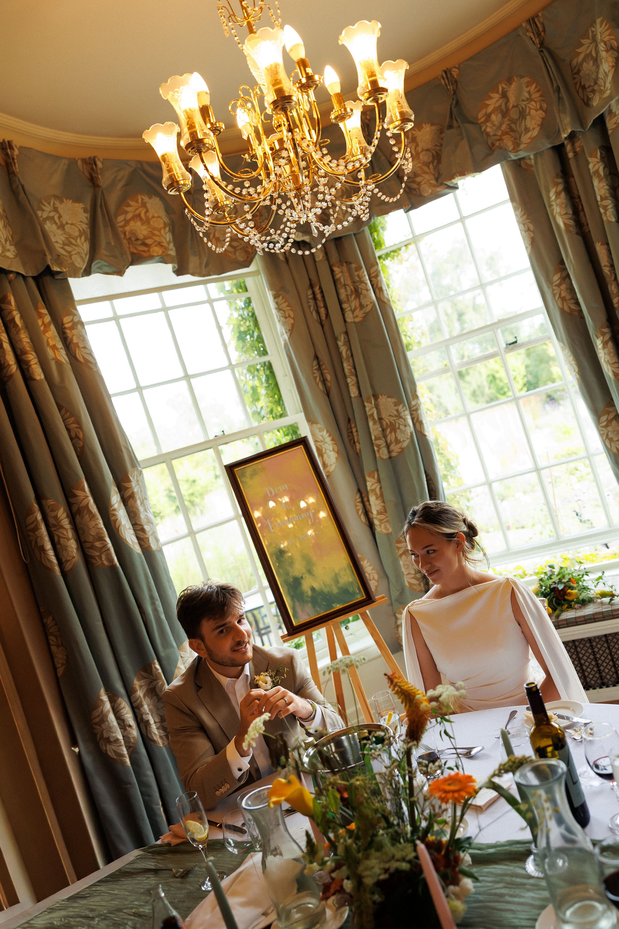 Elegant wedding couple dining under a crystal chandelier in a luxurious room with floral drapes and large windows.