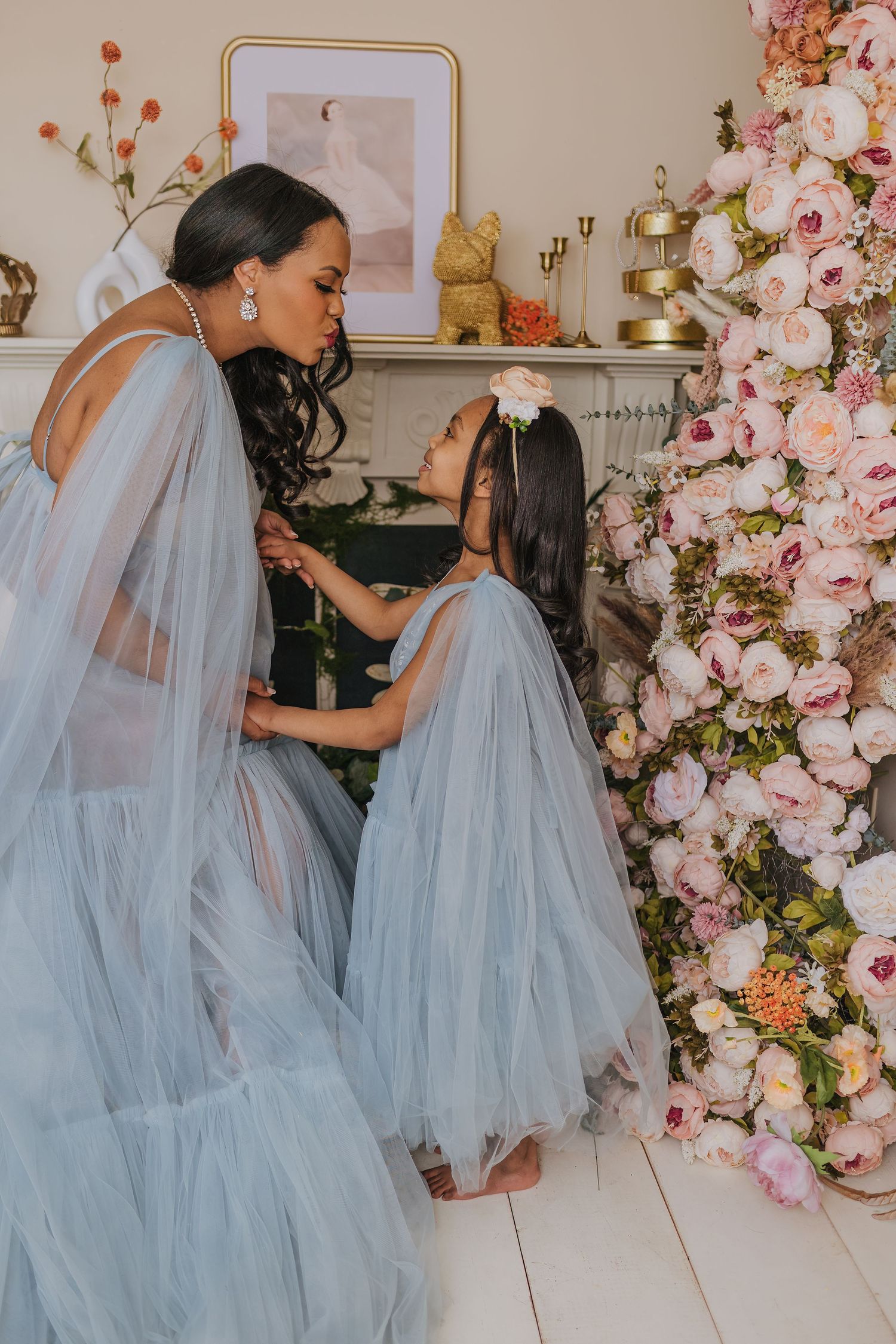 Two figures in ethereal blue tulle gowns share tender moment by pink floral arrangement.