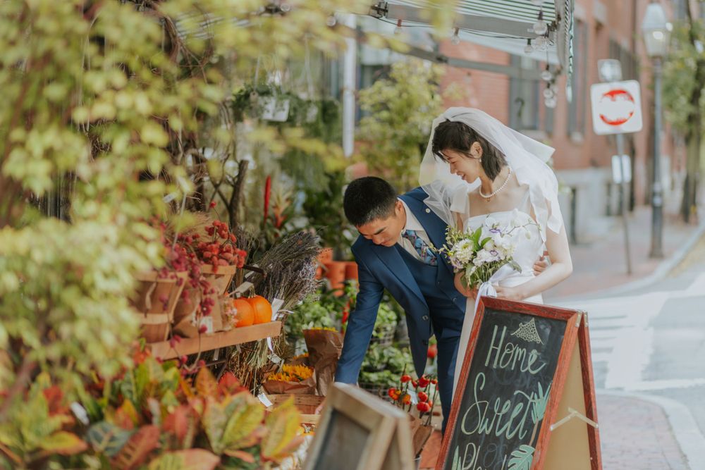 A couple in wedding attire browsing a flower shop display on a charming brick-lined street.