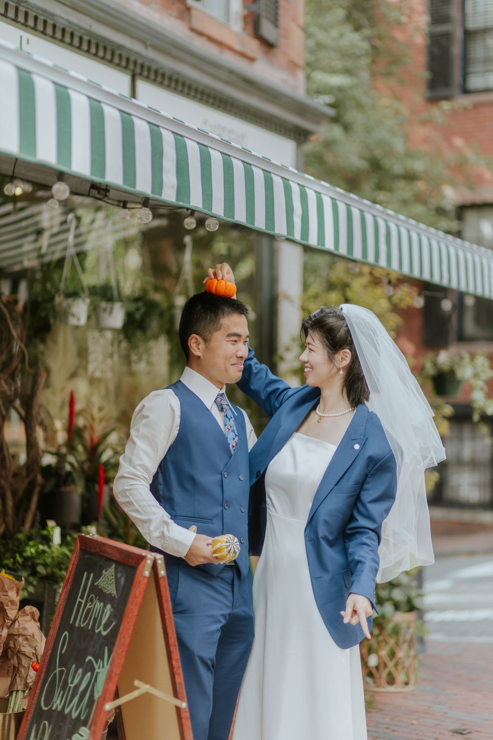 A couple in wedding attire poses in front of a striped awning and floral shop display.