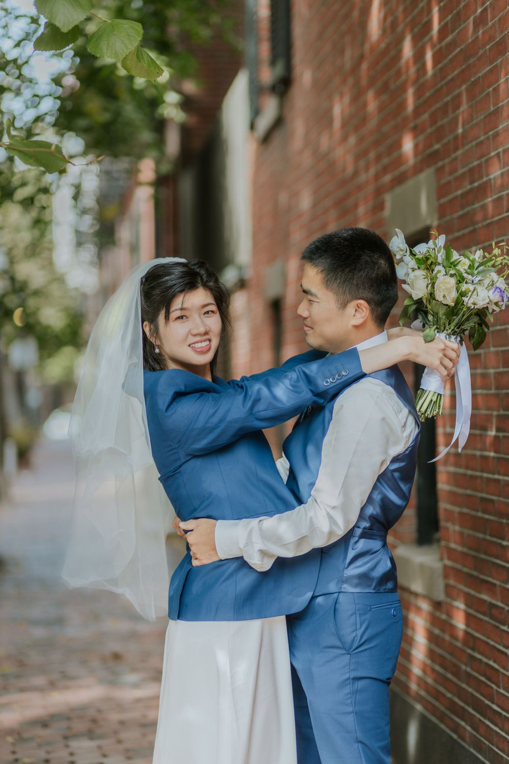 A couple in blue and white wedding attire embraces on a brick-lined street.