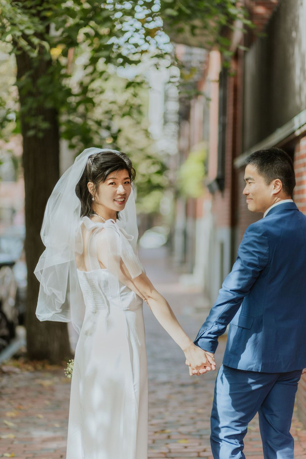 A couple in wedding attire walks hand in hand down a tree-lined brick street.