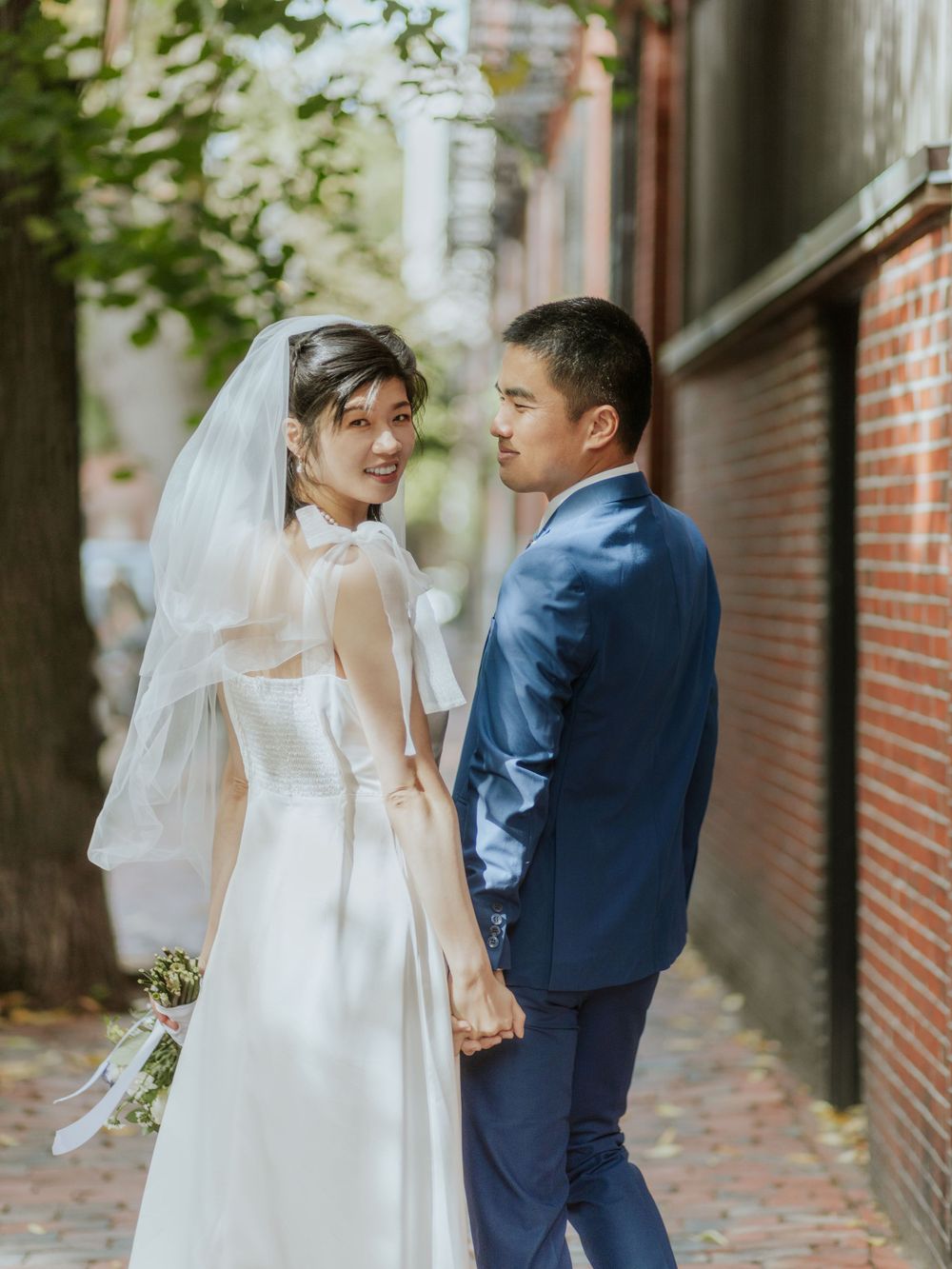 A couple in wedding attire looks back while walking down a brick alleyway.
