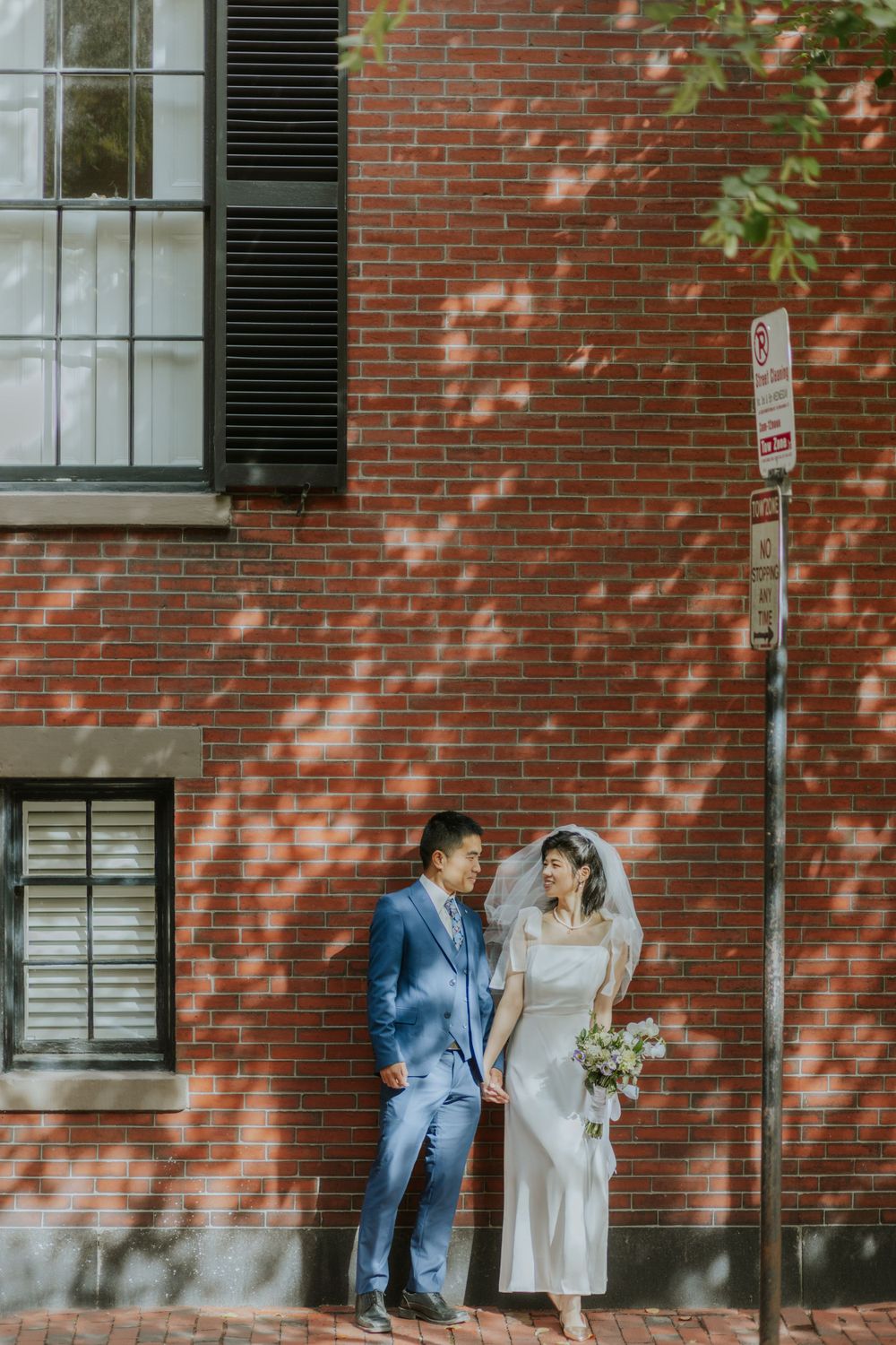 A couple in wedding attire poses against a red brick wall with dappled sunlight.