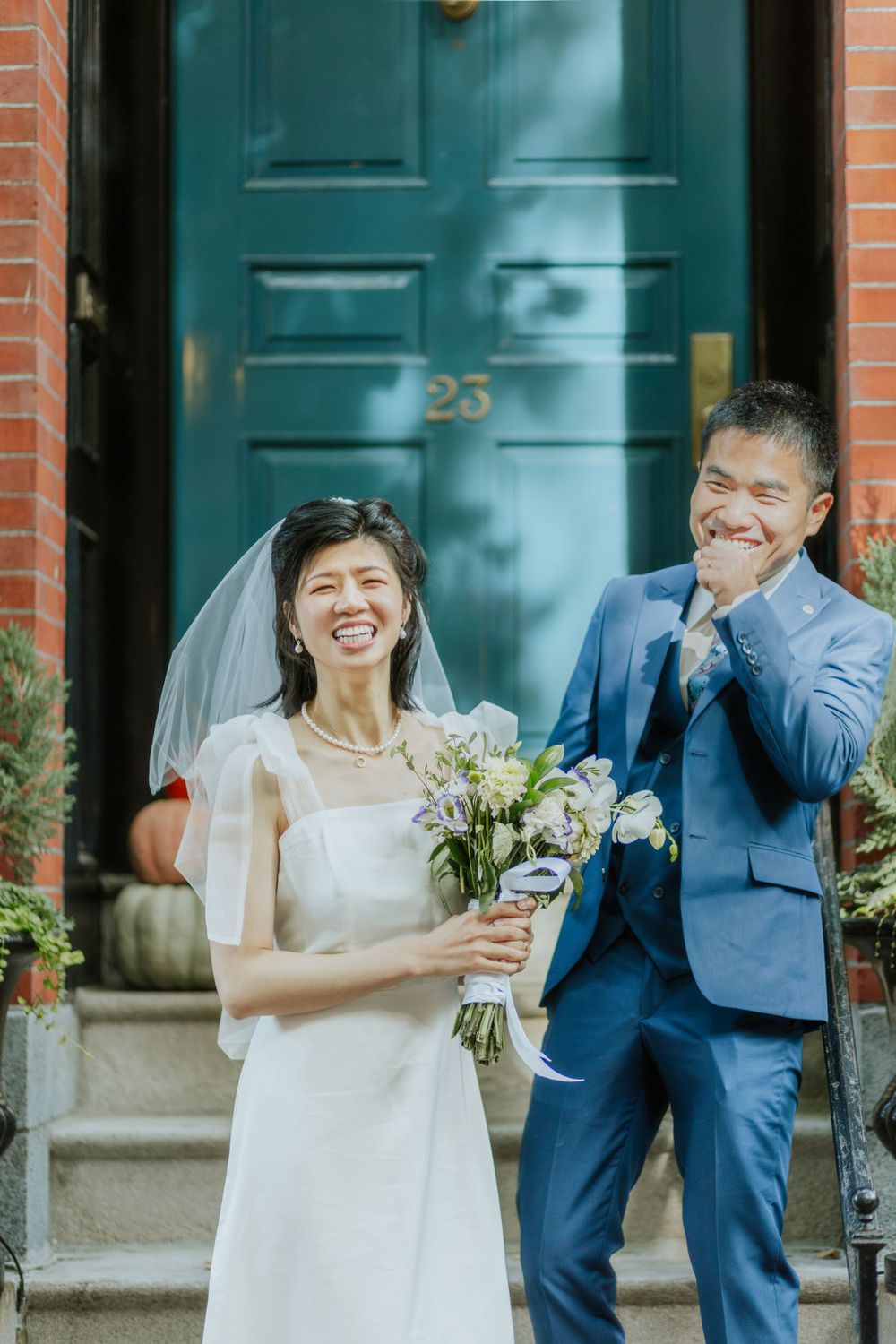A bride in white dress holding a bouquet shares a joyful moment with her groom in blue suit on brick steps.