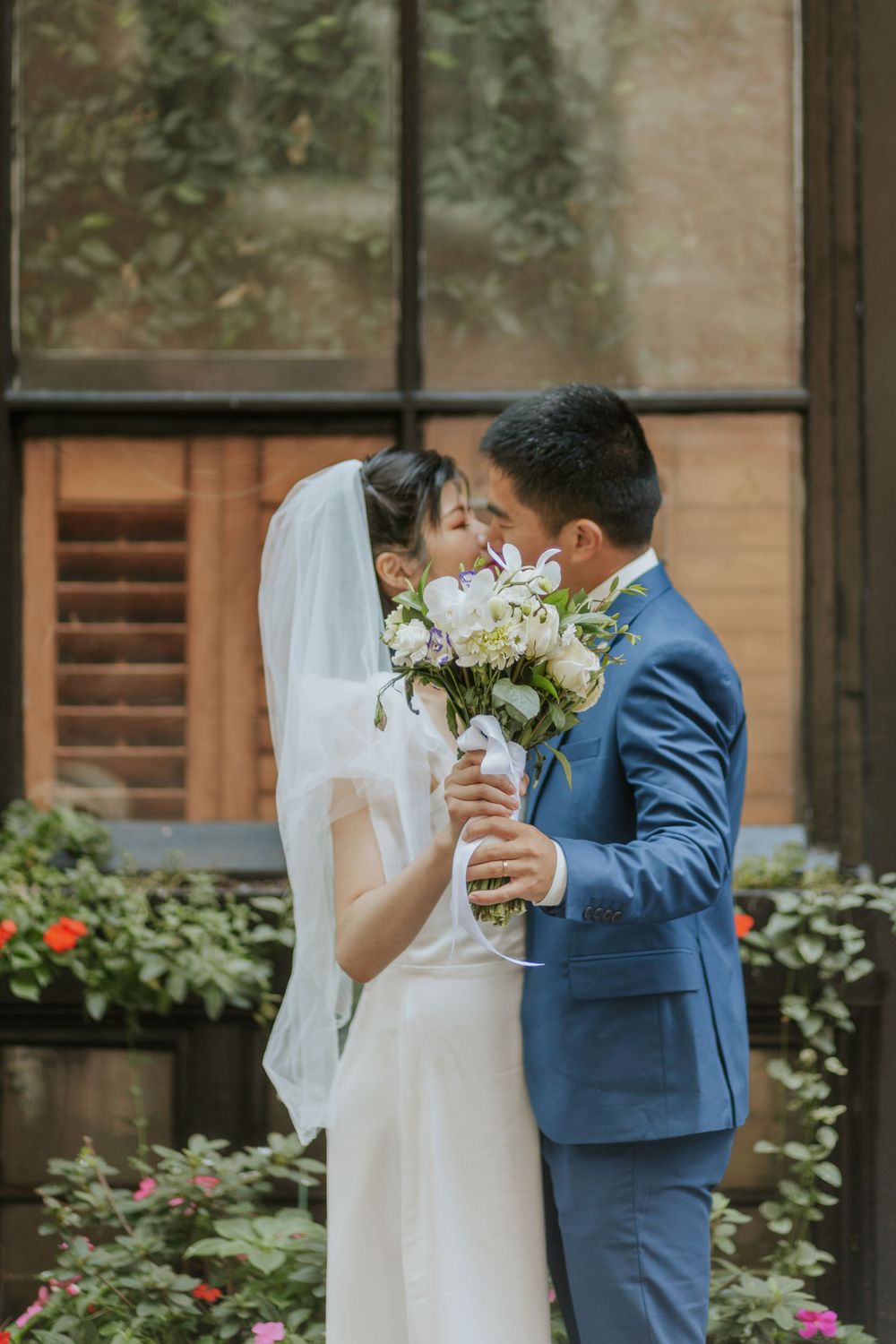 A romantic wedding photo of a couple sharing an intimate moment with white flowers against brick walls.