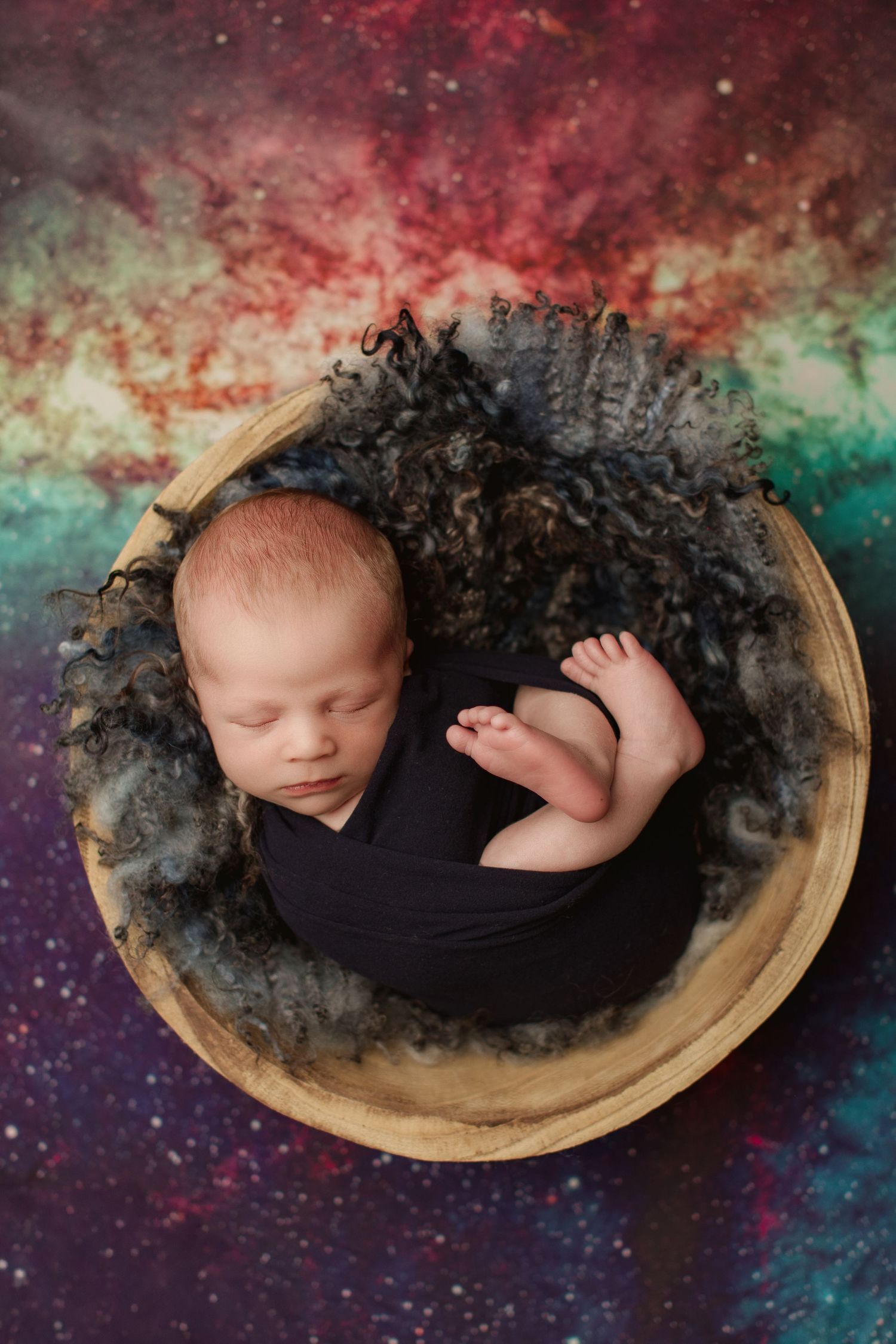 A sleeping newborn swaddled in black fabric rests peacefully in a wooden bowl against a colorful cosmic background.