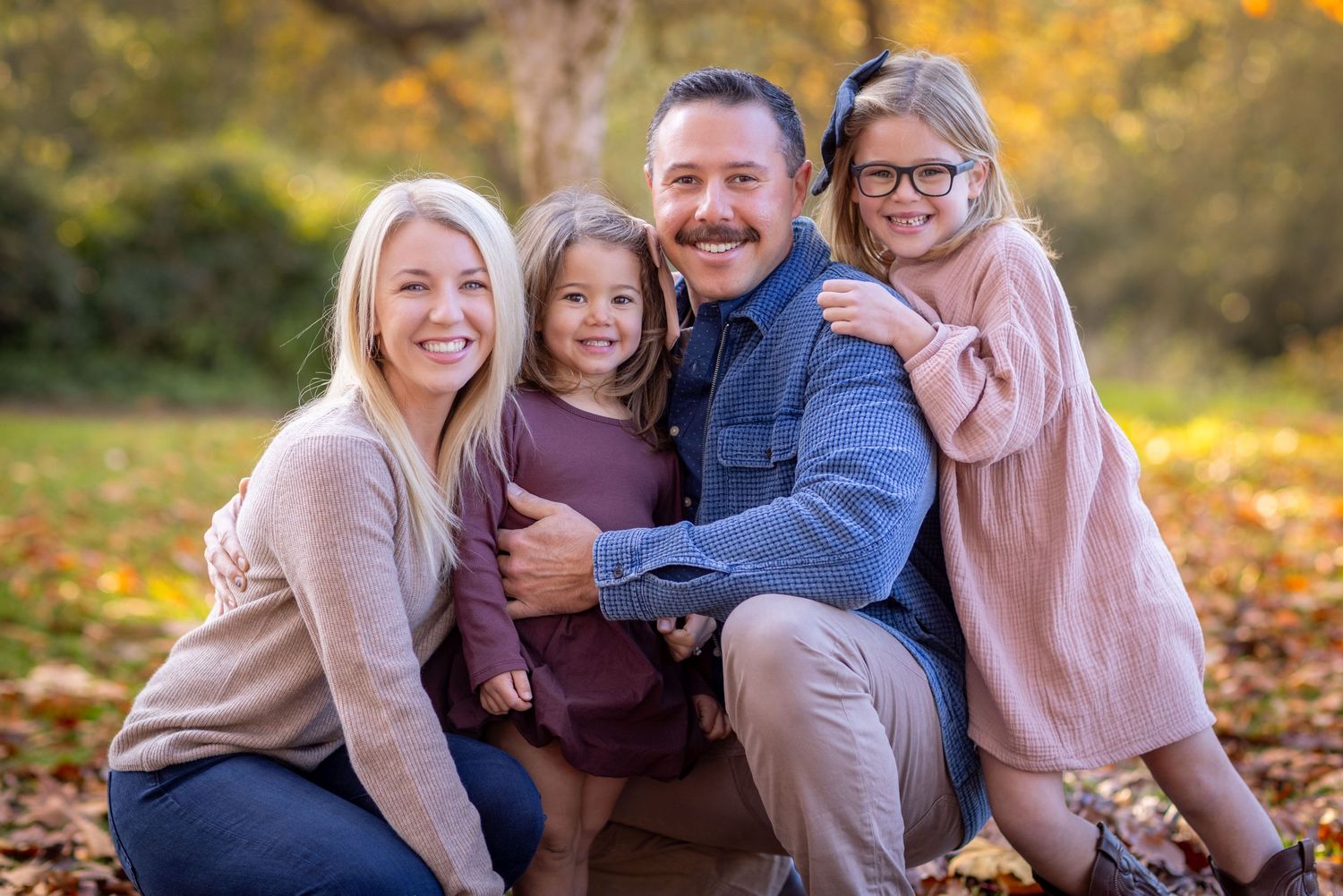 A family poses together outdoors during a fall photo session with golden sunlight and autumn leaves in the background.