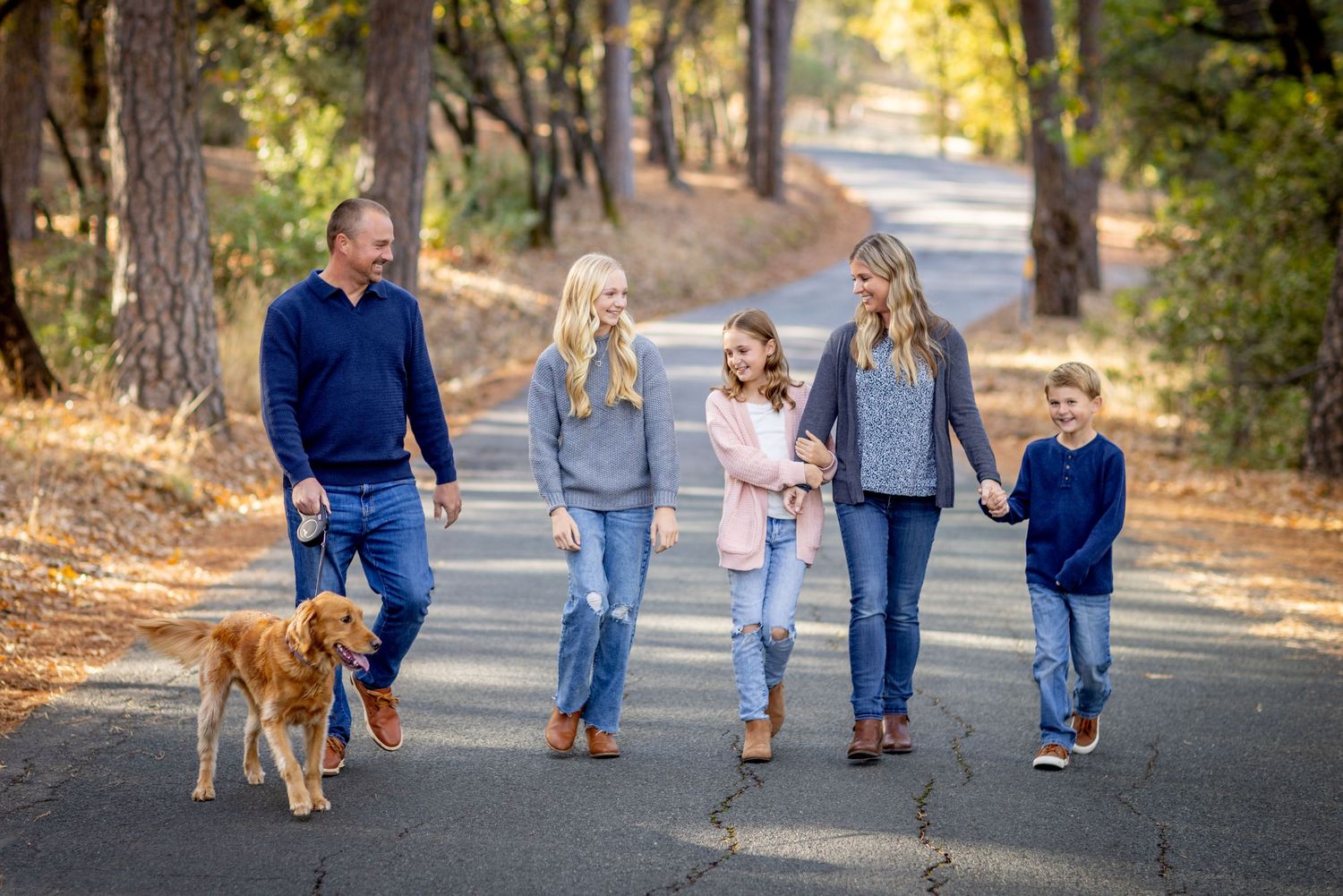 Family walking together with their golden retriever on a tree-lined path during a sunny autumn day.