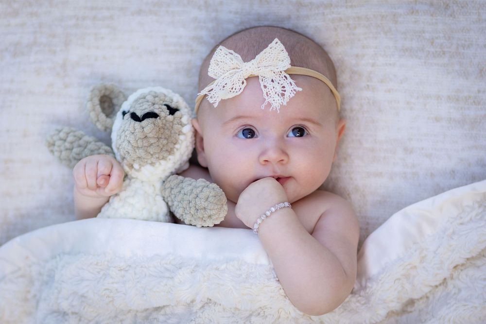 Sweet baby wearing delicate lace headband poses with soft plush lamb toy on white blanket.