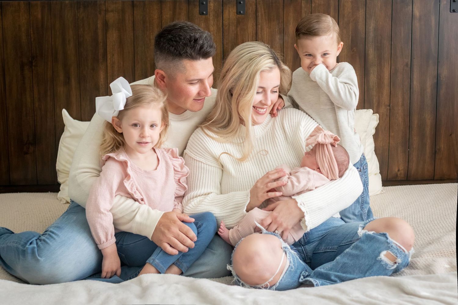 A family in matching cream and pink outfits sits together on a white bed against a wooden wall background.