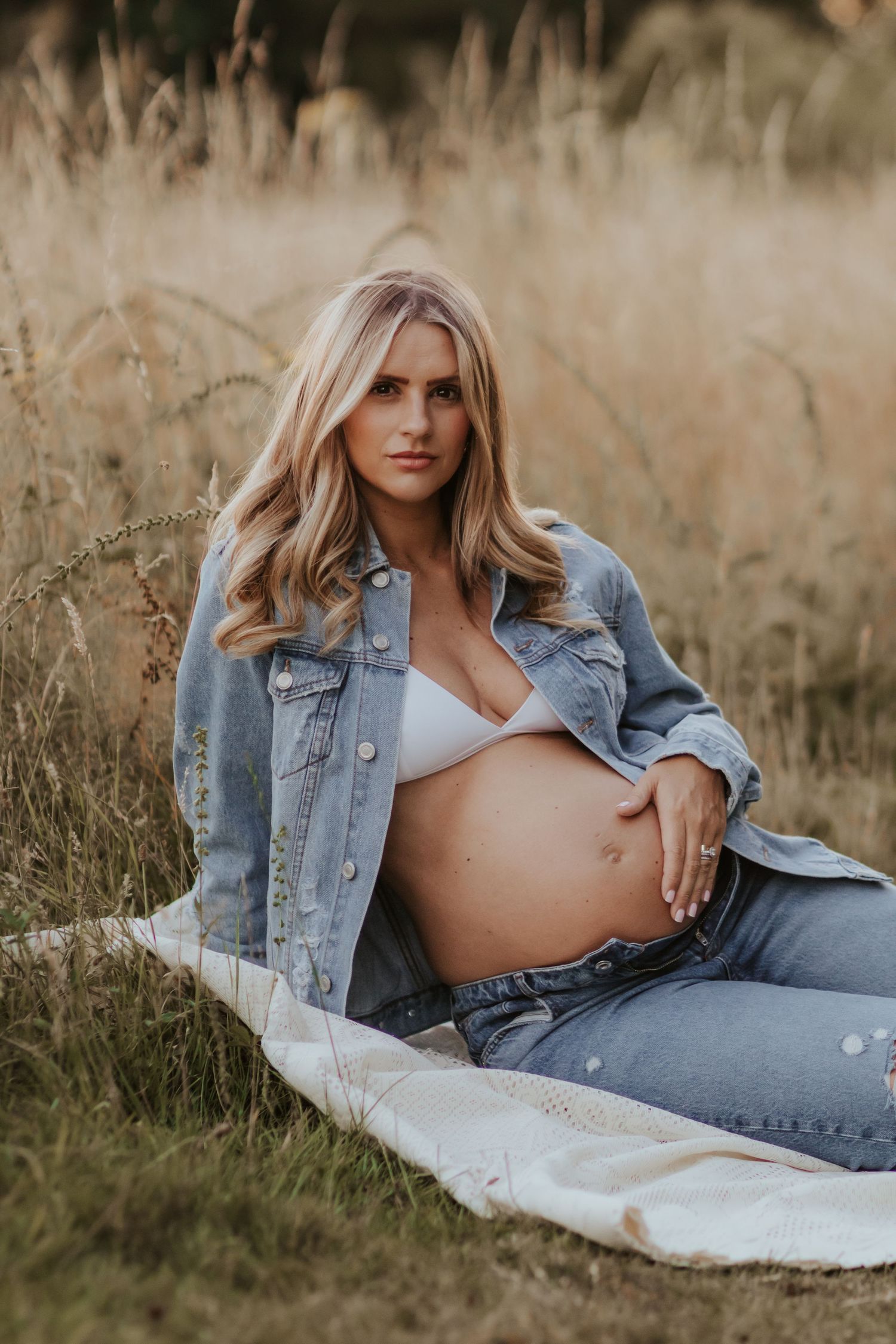 Maternity photoshoot in a field showing casual double denim style and bare belly against dried grass background.