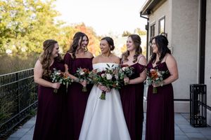 Bride in white gown laughing with four bridesmaids in burgundy dresses holding bouquets on an outdoor venue balcony.