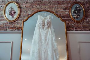Delicate lace wedding dress displayed in ornate gold mirror against brick wall.