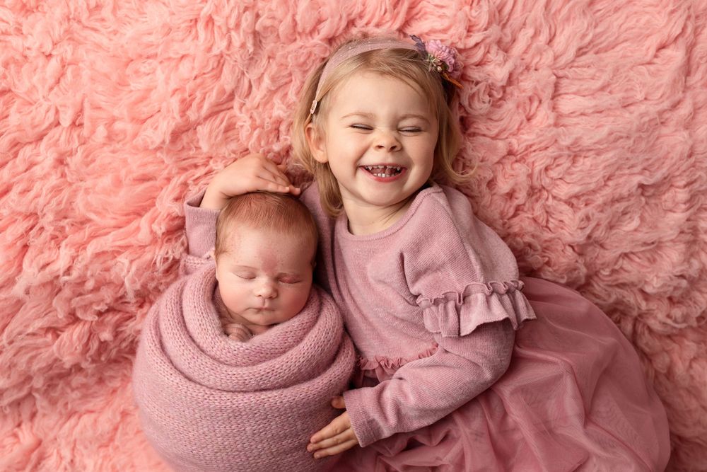 Two siblings posing together on pink fluffy blanket wearing matching pink outfits.