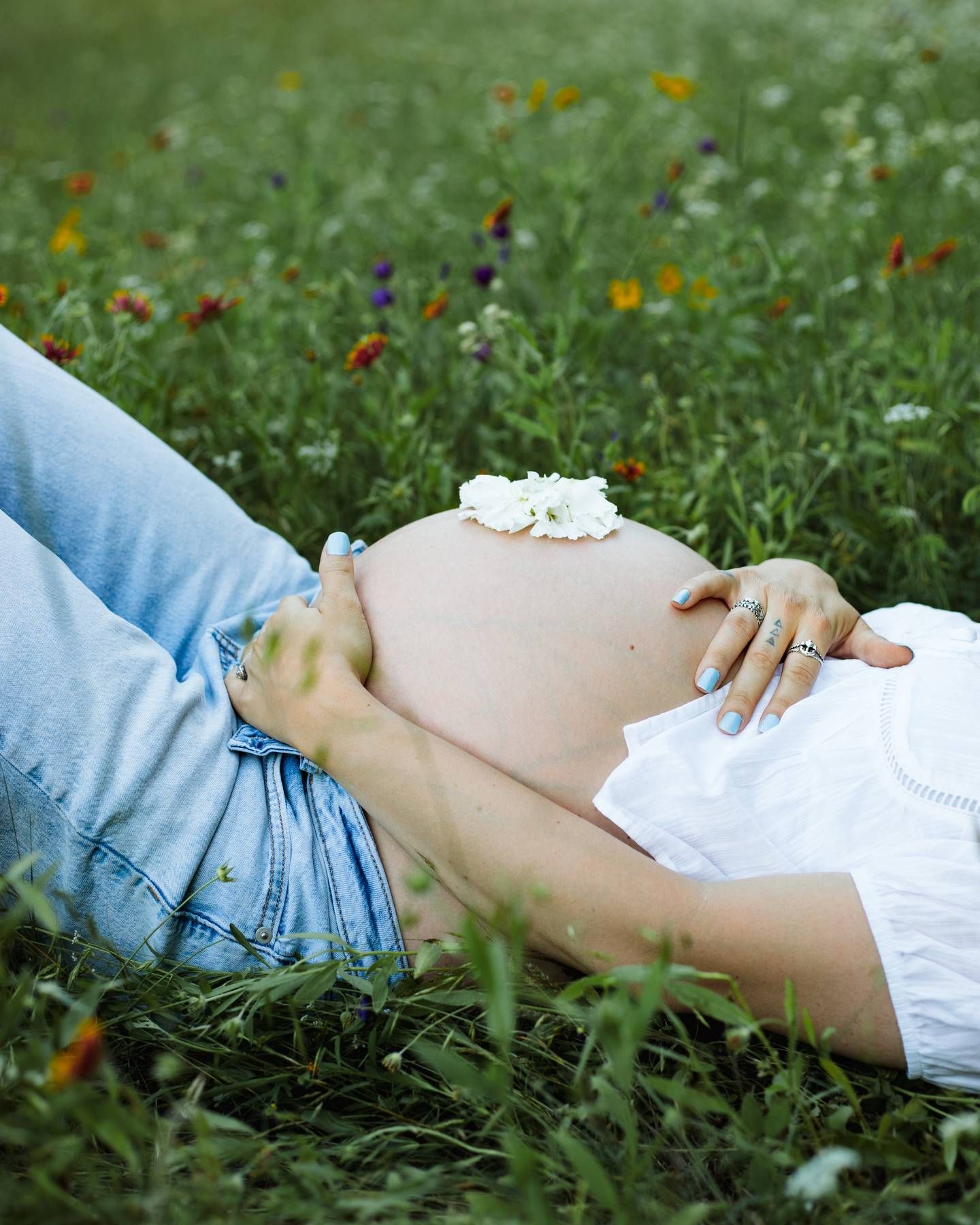 Pregnant woman in jeans and a white top rests on a wildflower meadow with a white flower on her baby bump.