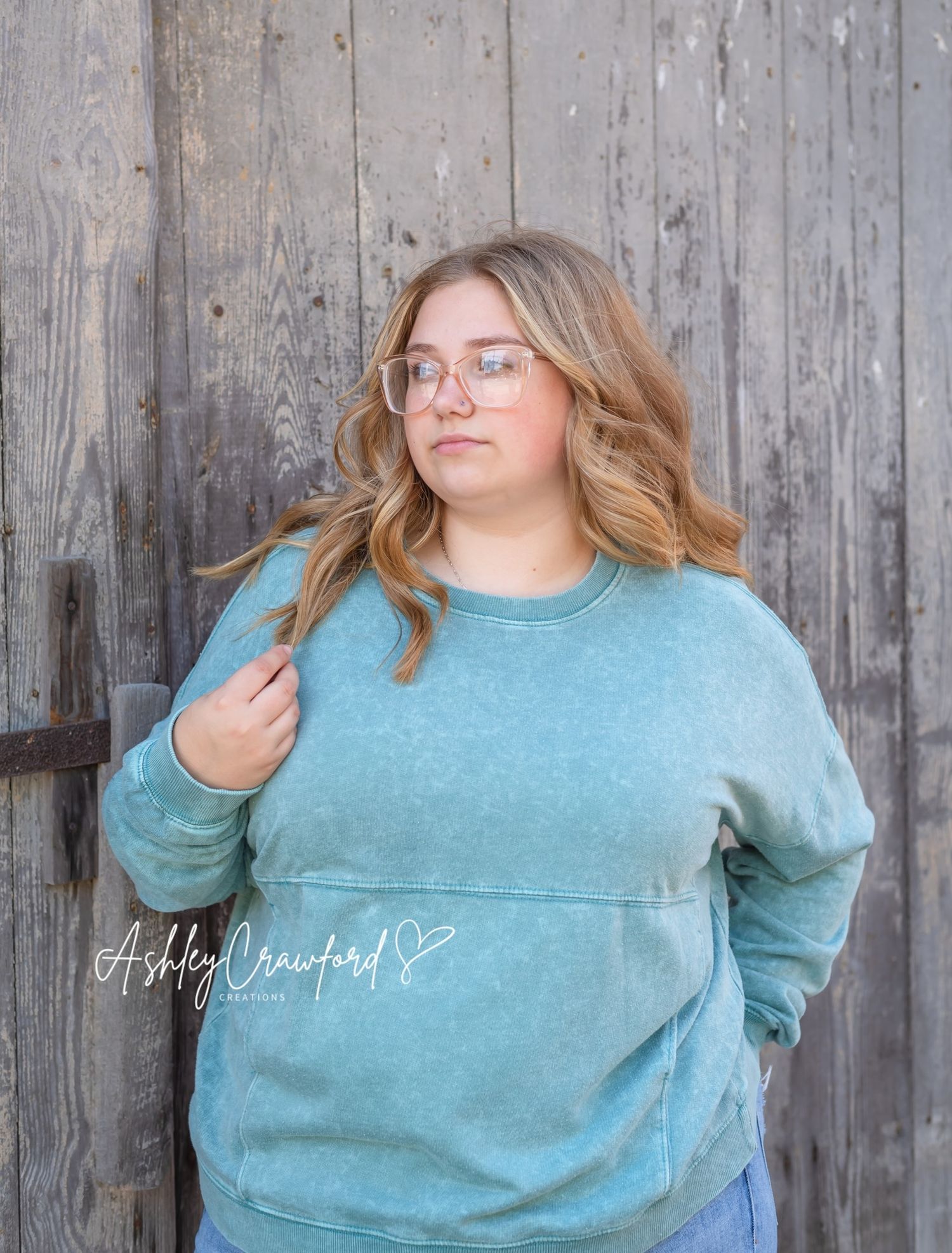 Portrait of woman in teal sweatshirt posing against weathered wooden wall backdrop.