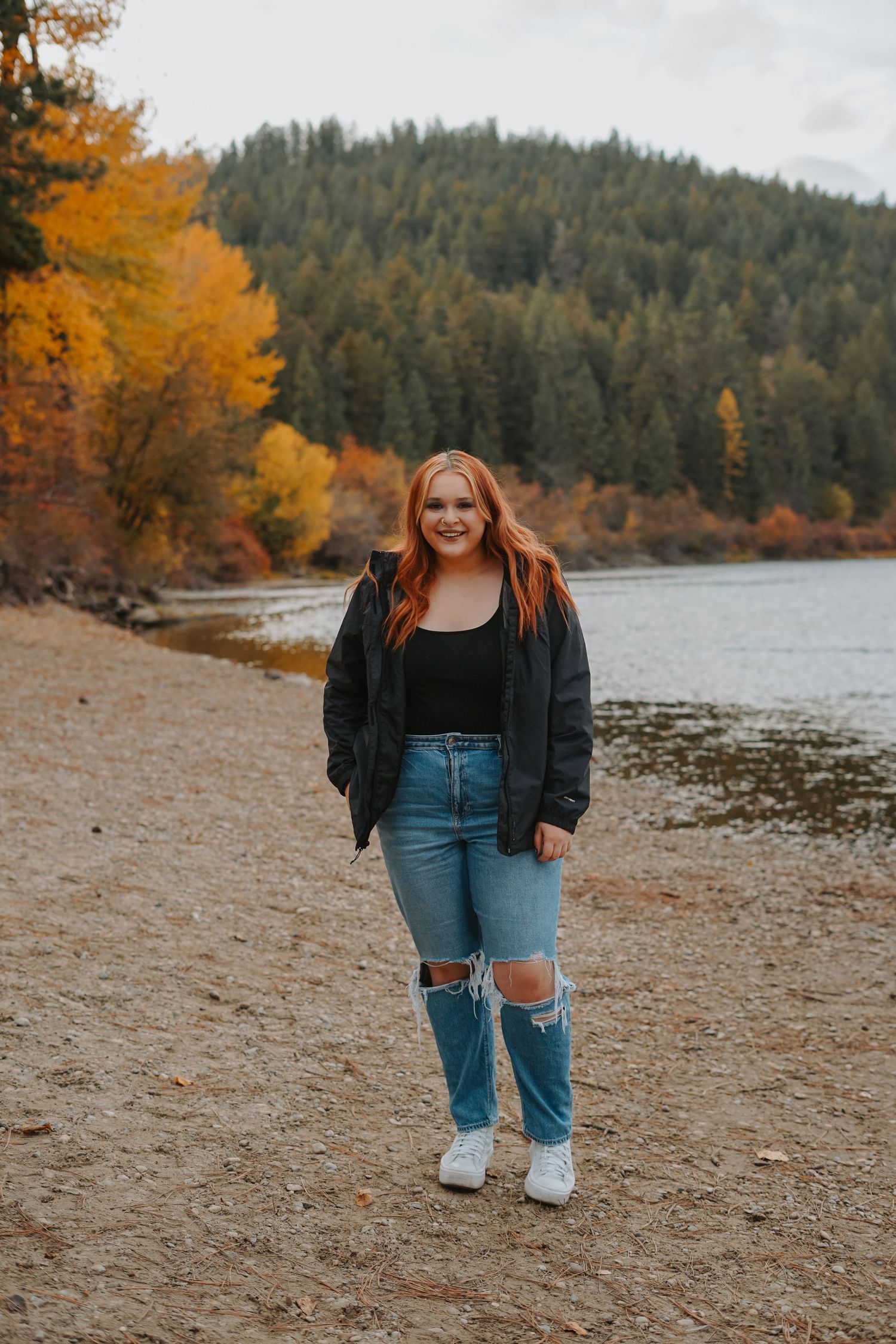Person stands on autumn lakeshore with colorful fall foliage and evergreen forest backdrop.