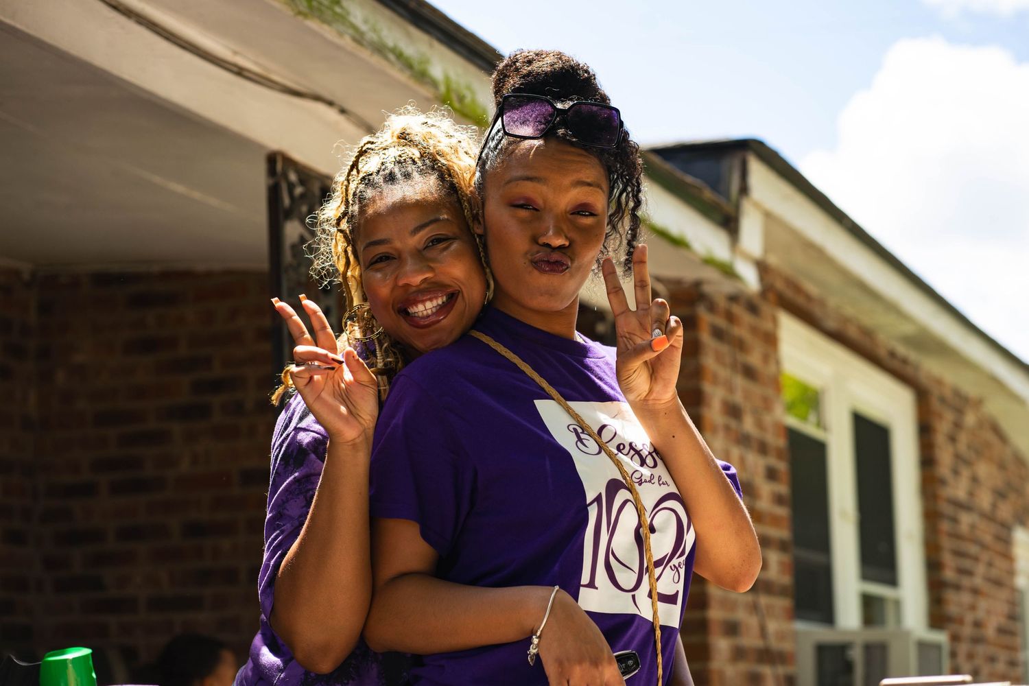Two people in purple t-shirts making peace signs and smiling outdoors.