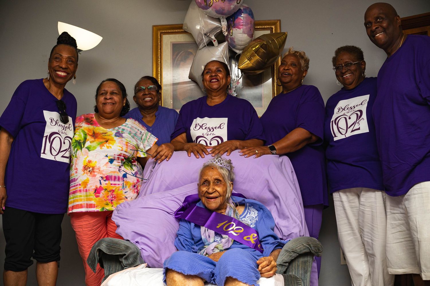 A group wearing matching purple shirts gathers around a bed with celebratory balloons and decorations.