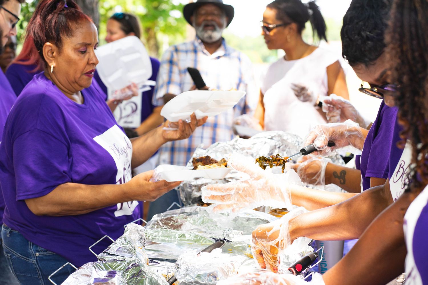 People in purple shirts preparing and serving food at an outdoor gathering.