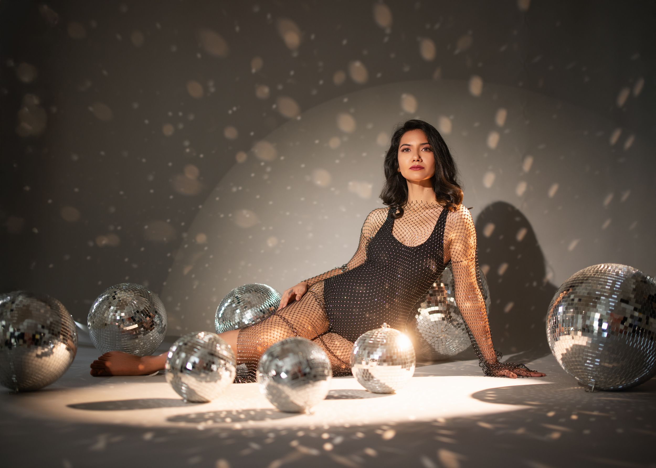 Model in a black dress poses with scattered disco balls and dramatic lighting creating a glamorous studio atmosphere.