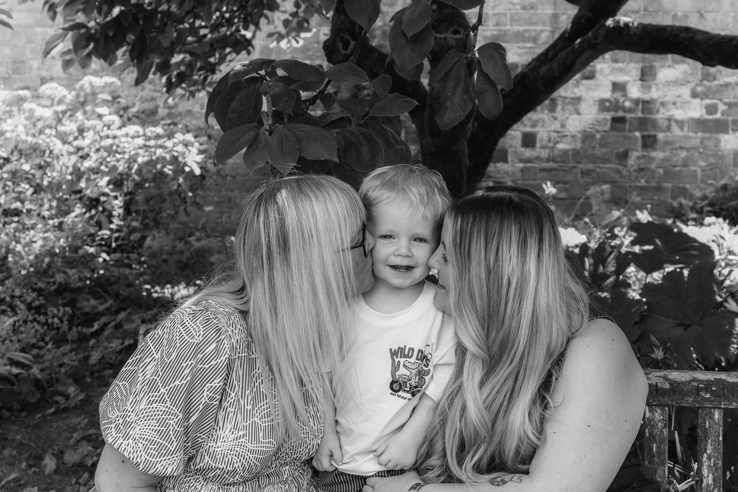 Black and white photo of grandmother and daughter kissing a smiling toddler boy on the cheeks during outdoor family photoshoot