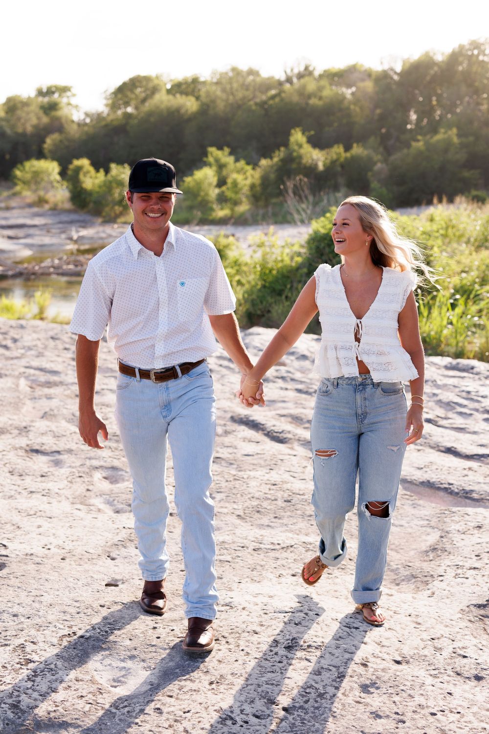 Couple in coordinated white and denim outfits walking hand in hand on sandy beach.