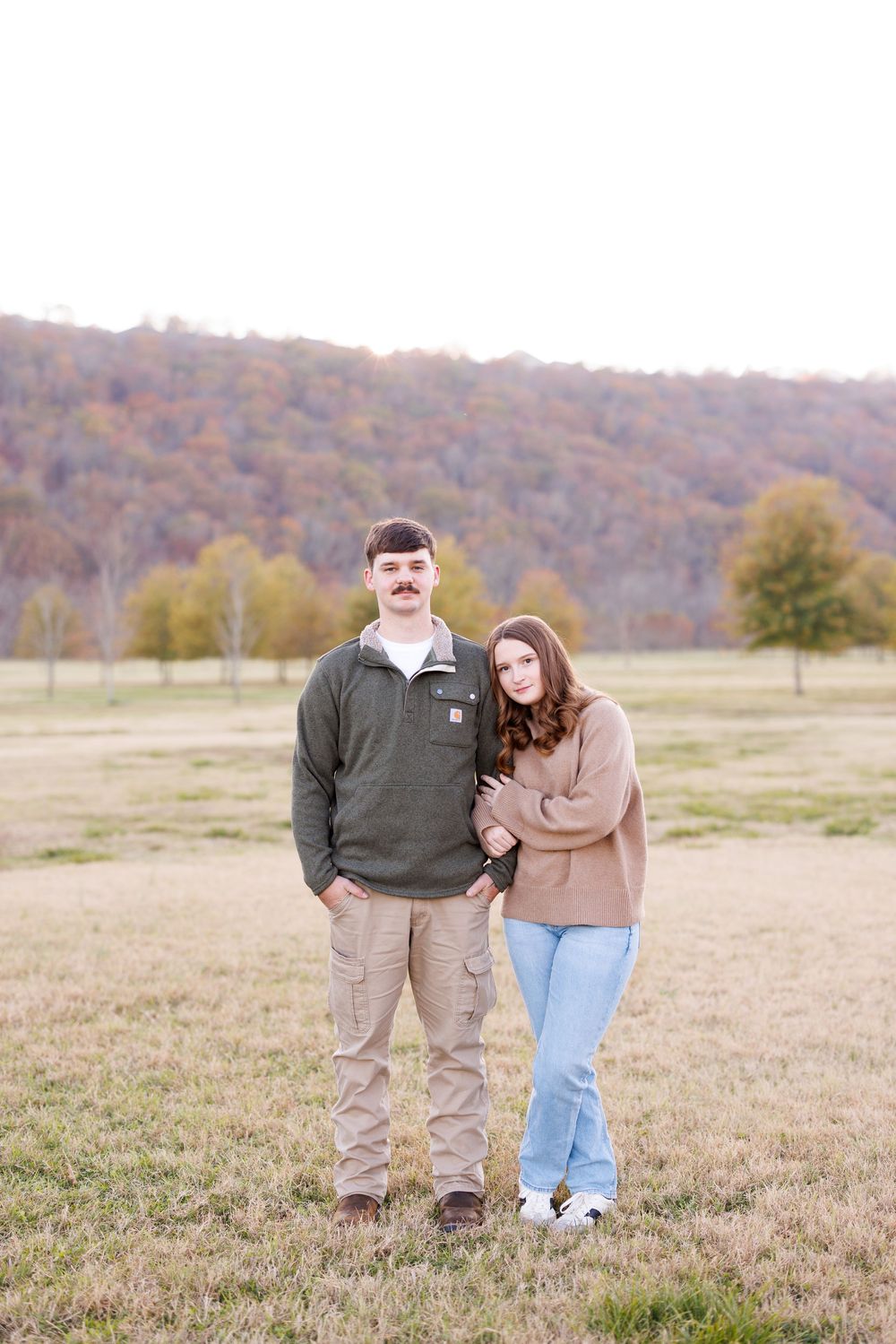 A couple laughs together in a field with autumn hills in the background.