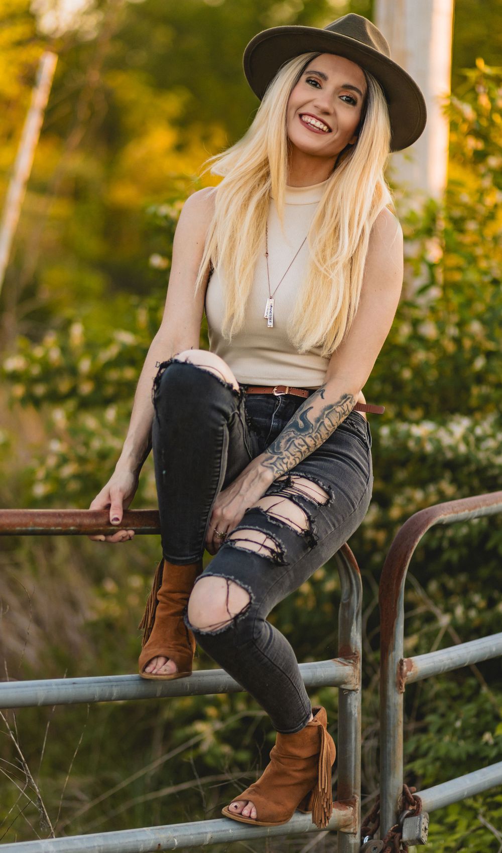 Woman in fedora and distressed jeans sitting on metal railing during golden hour.