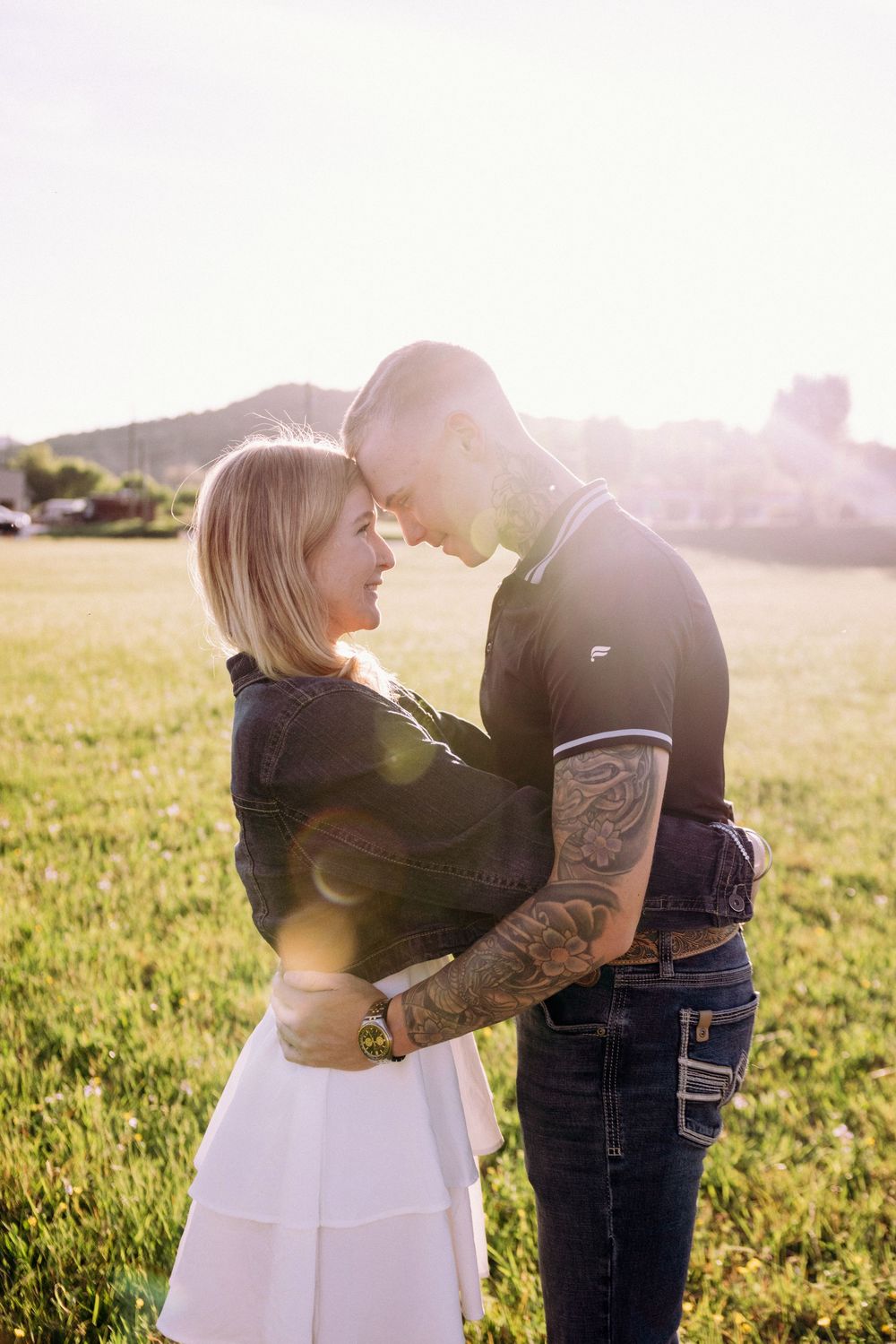 Couple embracing in a sunny green field, woman in white skirt and man with tattoos in navy polo, backlit by golden sunlight.