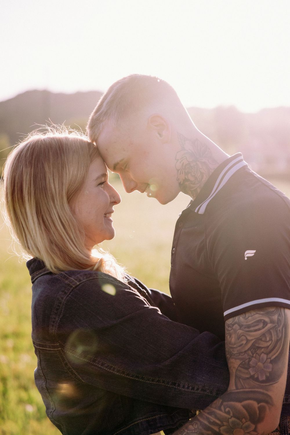 A couple with tattoos sharing a romantic moment outdoors, smiling at each other in golden sunlight.