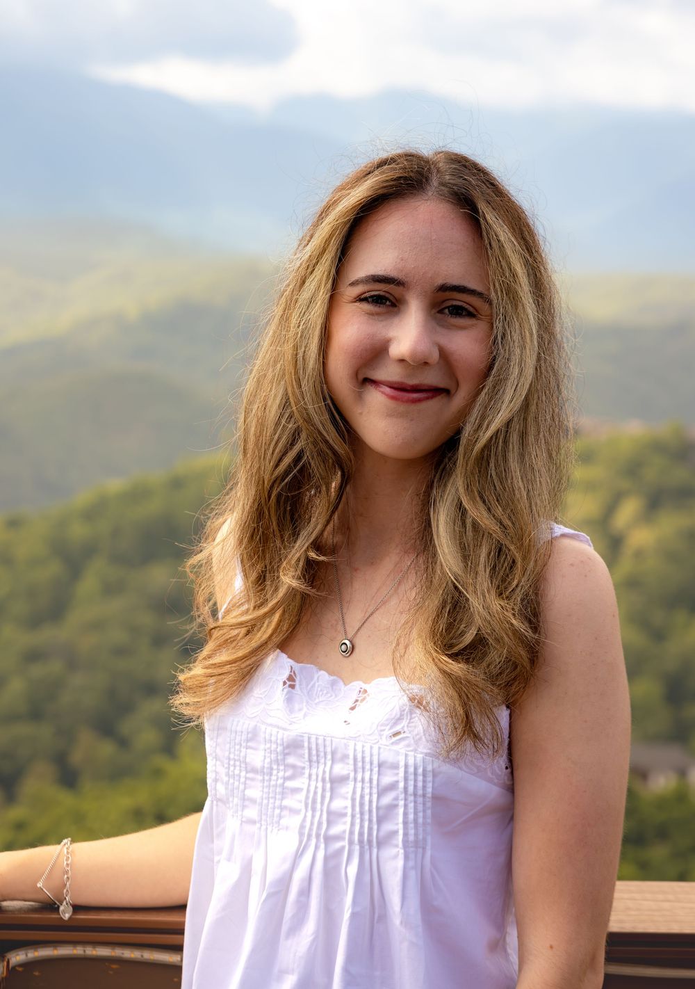 Young woman with blonde hair smiling on a deck with scenic green mountain landscape in the background.
