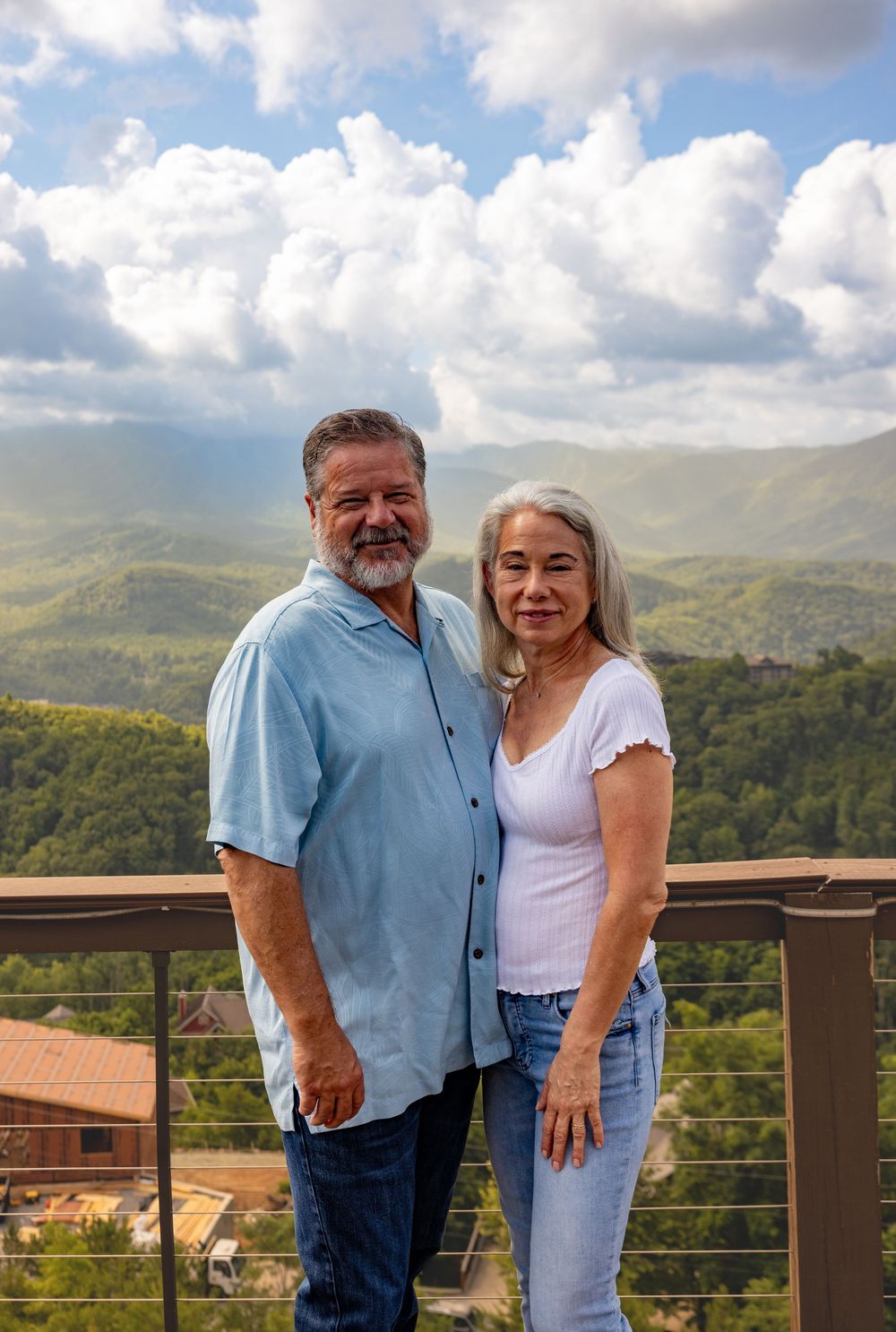 A smiling couple poses on a balcony with scenic green mountain views and cloudy blue sky backdrop.
