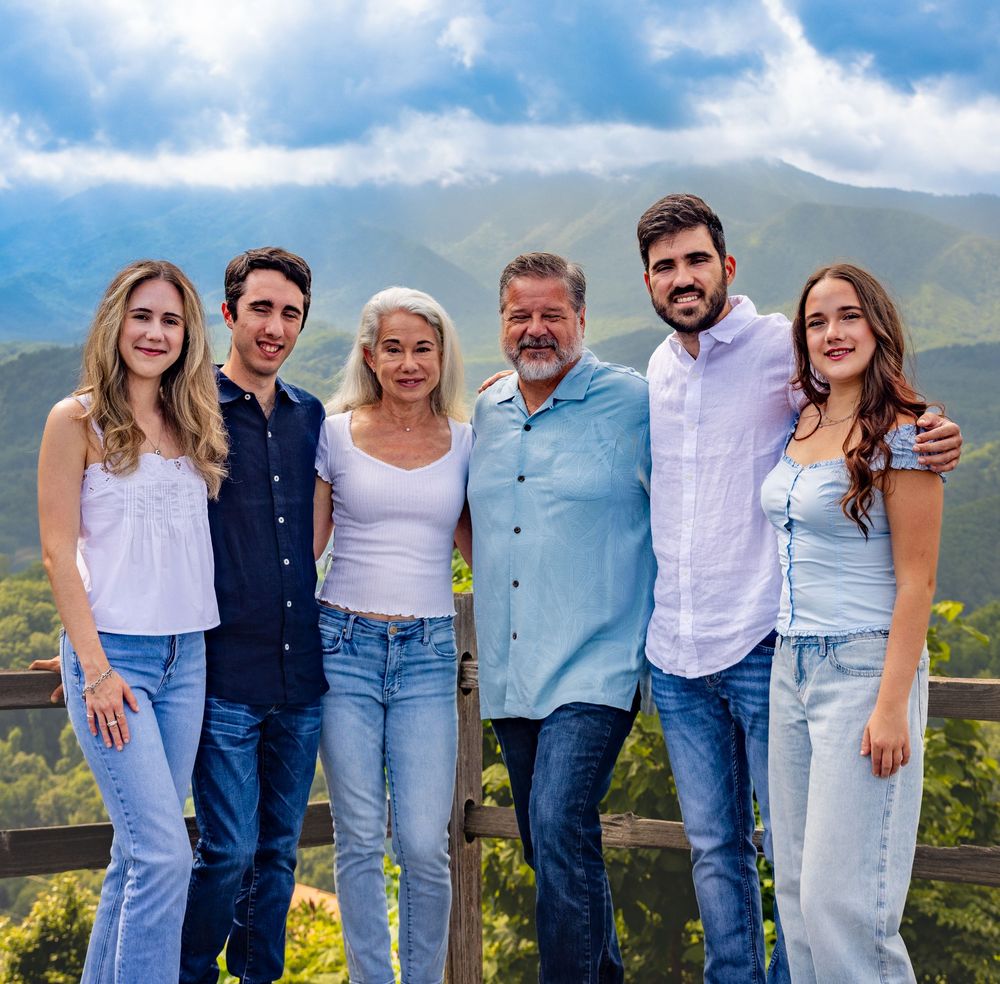 Family of six smiling together on a scenic mountain overlook under a cloudy sky.